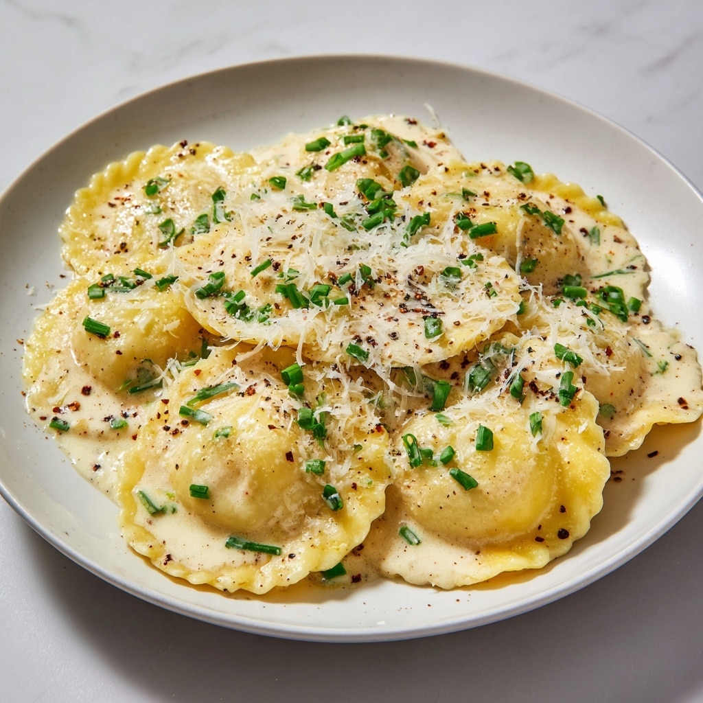 A close-up view of a white plate filled with round ravioli pieces sitting in creamy white sauce, each ravioli having a smooth yellow surface. On top, there is a layer of finely grated white cheese scattered over the ravioli along with small green chive pieces spread evenly across the dish. Black pepper flakes are lightly sprinkled around, adding small dark specks on the sauce and pasta. The white marbled background adds a clean and bright feel to the image. Photo taken with an iphone --ar 4:5 --v 7