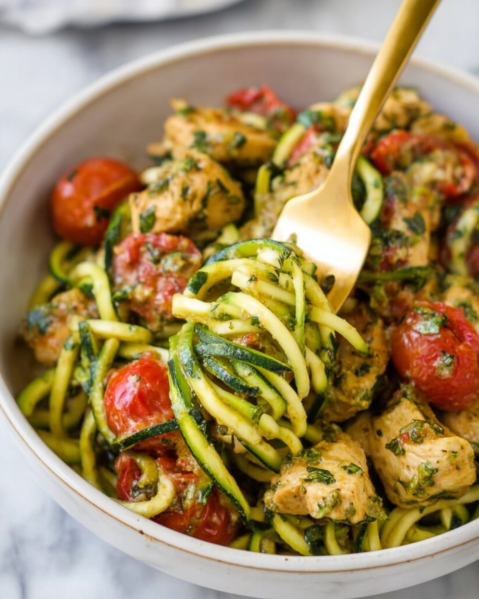 A close-up view of a white bowl filled with zucchini noodles mixed with green pesto sauce, light brown cooked chicken pieces, and red cherry tomato halves. The zucchini noodles are thin, spiral-shaped, and glossy, forming the base layer. Scattered on top are chunky pieces of tender chicken coated in a green pesto herb mixture. Bright red cherry tomatoes add bursts of color throughout the dish. A gold fork is stuck into the bowl, lifting a small bundle of noodles, chicken, and tomato. The background is a white marbled surface. photo taken with an iphone --ar 4:5 --v 7