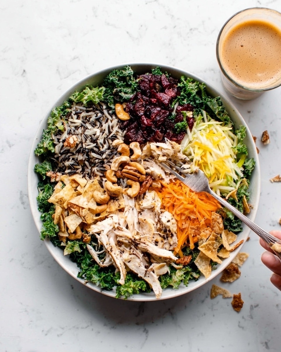 A white bowl is filled with a colorful layered salad arranged in six sections on a white marbled surface. Starting from the top left and moving clockwise, there are dark green kale leaves, a mix of wild rice with black and brown grains, light cooked white shredded chicken, golden cashews, shredded pale yellow cheese, and bright chopped red vegetables with crispy light brown chips. A woman's hand is lifting some of the salad with a fork from the edge of the bowl. A glass of iced coffee is visible in the top right corner. photo taken with an iphone --ar 4:5 --v 7