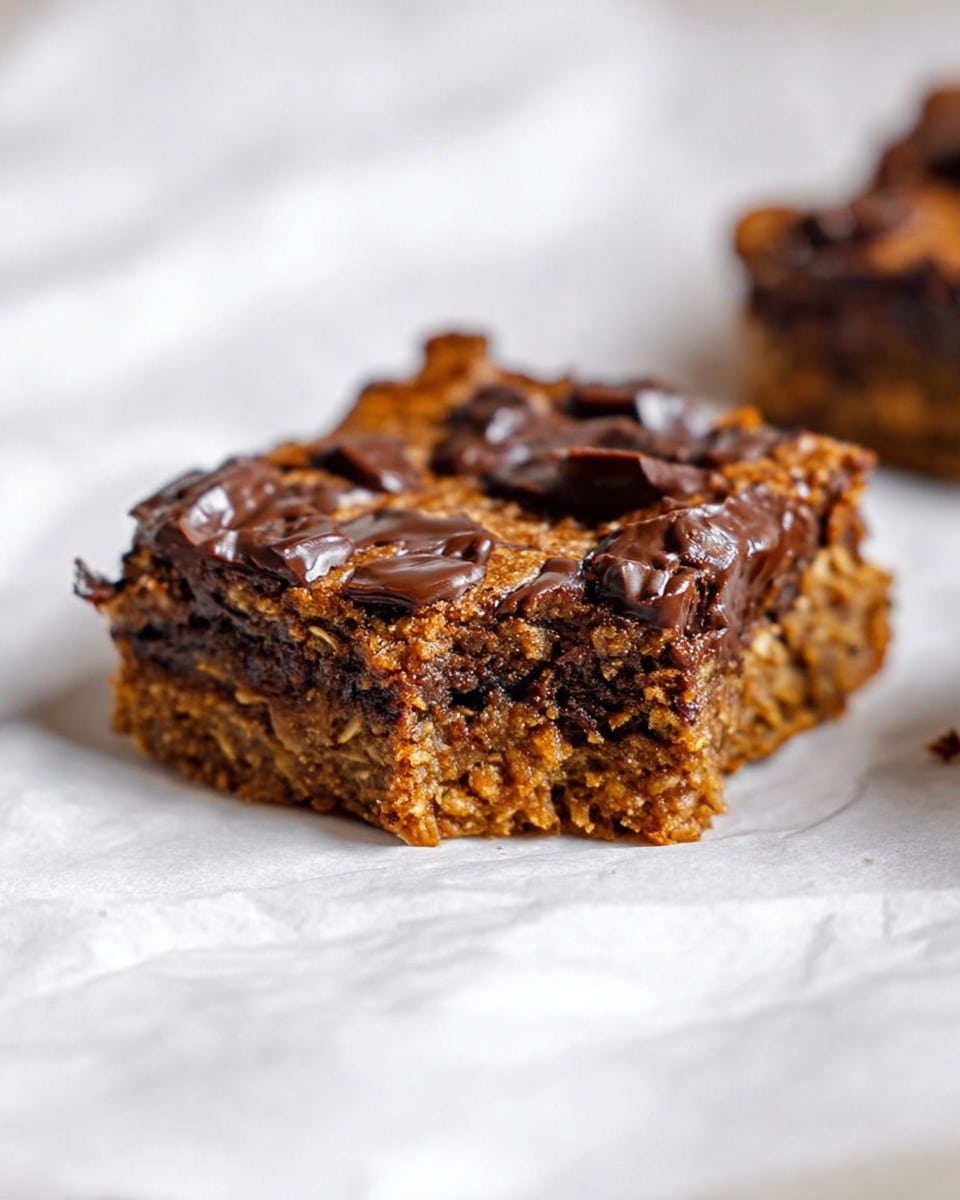 A thick, square piece of baked dessert with a slightly crumbly texture sits on white parchment paper over a white marbled surface. The dessert has two visible layers made of a brown, oat-filled base and middle, with melty, dark chocolate chunks generously spread inside and on top. The top layer shows scattered, glossy dark chocolate pieces partially melted into the soft, dense oat mixture. photo taken with an iphone --ar 4:5 --v 7