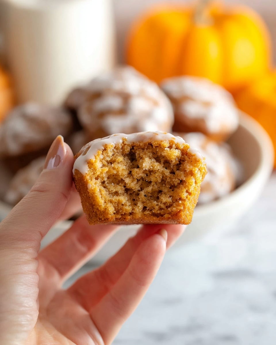 A white bowl filled with many round pumpkin spice donut holes covered in a light brown glaze, stacked in a heap. The donut holes have a rough, slightly crumbly texture with the glaze giving a shiny, smooth coat on top. Each donut hole shows small sugar bits and pumpkin spice specks, making the surface lumpy and uneven. In the background, there is a small orange pumpkin and white marbled surface below the bowl. The lighting is soft and natural, highlighting the warm colors of the donuts. photo taken with an iphone --ar 4:5 --v 7
