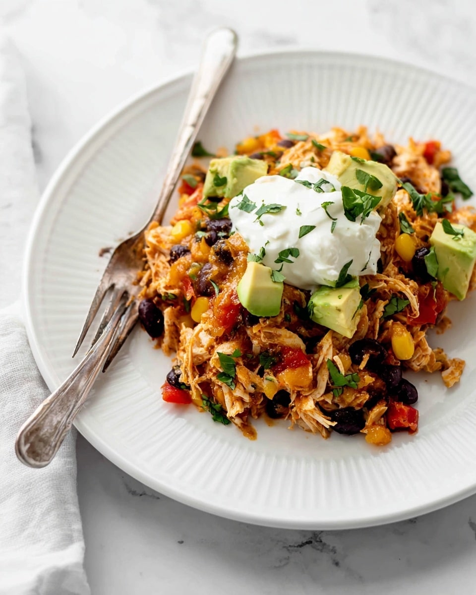 The dish is served on a white plate with ridged edges, set on a white marbled surface. It has a base layer of shredded chicken mixed with black beans, corn, diced red bell peppers, and fresh herbs, giving a mix of light brown, black, yellow, red, and green colors. Scattered on top are small pieces of diced avocado, adding a fresh light green color. On the very top, there is a dollop of thick, white sour cream. A silver fork rests on the left side of the plate, partly touching the food. Photo taken with an iphone --ar 4:5 --v 7