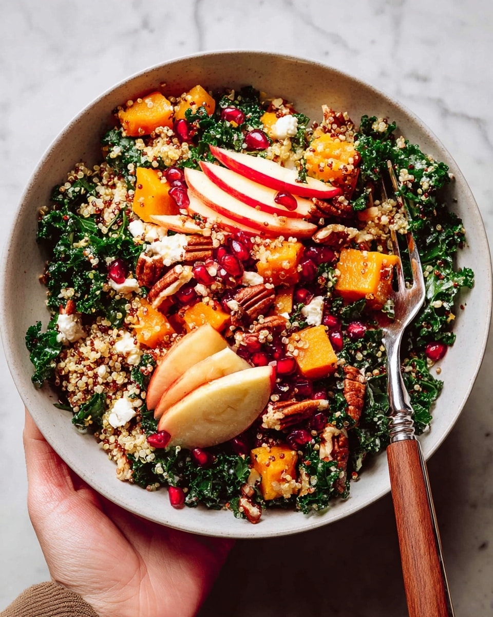A white bowl filled with a colorful salad showing three main layers: the base layer is dark green kale leaves with a rough texture, mixed with light beige quinoa grains scattered evenly; the middle layer features bright orange roasted squash cubes alongside thin, curved slices of red apple with white flesh; the top layer is sprinkled with small bright red pomegranate seeds and broken pieces of white feta cheese, plus glossy dark brown pecans, all mixed evenly. A silver fork with a wooden handle rests inside the bowl on the right side. A woman's hand is holding the bowl at the bottom left corner against a white marbled textured surface. Photo taken with an iphone --ar 4:5 --v 7