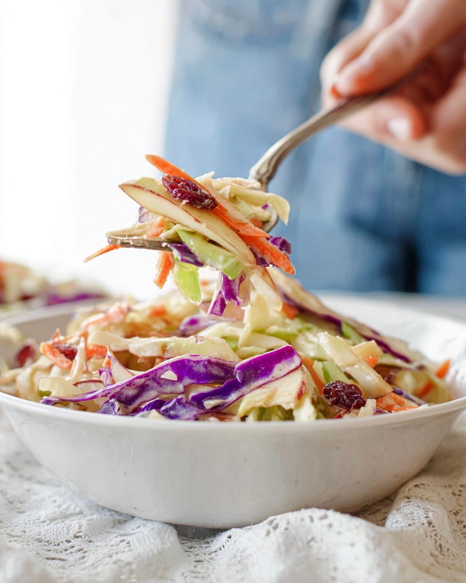 A close-up of a colorful coleslaw salad showing two layers in a white bowl. The base layer is shredded green and purple cabbage with thin orange carrot strips scattered throughout. Mixed among the vegetables are small pieces of pale apple slices and reddish dried cranberries, adding spots of bright red and green. The salad looks creamy with a light coating of dressing that gives it a soft texture and slight shine. A woman's hand holds a fork lifting a bite, emphasizing the mix of colors and textures in the salad against a blurred background. The white bowl sits on a soft white lace fabric over a white marbled surface. photo taken with an iphone --ar 4:5 --v 7