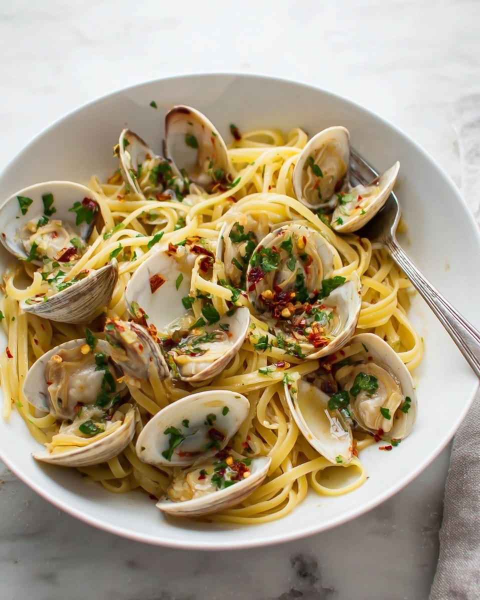 A large white pot with dark blue handles holds a dish made of two main layers: the bottom layer is light yellow cooked spaghetti pasta that looks soft and slightly glossy, and the top layer consists of open clam shells with pale grayish-white exteriors and peachy-beige clam meat inside, scattered evenly over the pasta. Bright green chopped parsley is sprinkled over the whole dish, adding small pops of color, and a small white bowl with more chopped parsley sits nearby on a white marbled surface. photo taken with an iphone --ar 4:5 --v 7