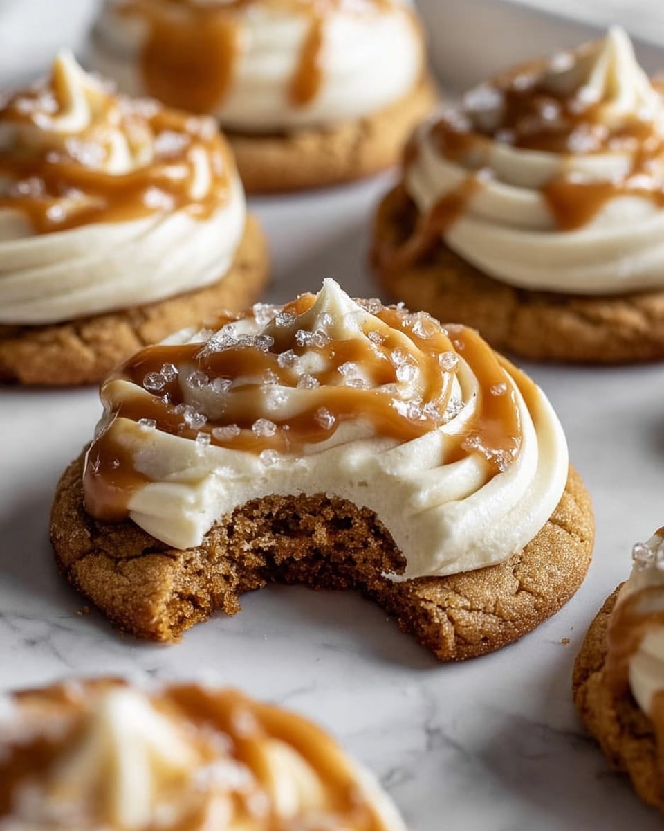 The image shows several round cookies on a white marbled surface, each with a golden-brown textured base. On top of the cookie base, there is a thick layer of smooth, creamy white frosting. Above this frosting, a swirl of the same creamy white frosting forms a small peak at the center. A glossy caramel sauce is drizzled generously over the frosting in an irregular pattern, and some coarse sea salt crystals are sprinkled on top, adding a sparkly texture. One cookie in the foreground has a bite taken out, revealing the soft, chewy inside and crumbly texture at the edge. Photo taken with an iphone --ar 4:5 --v 7