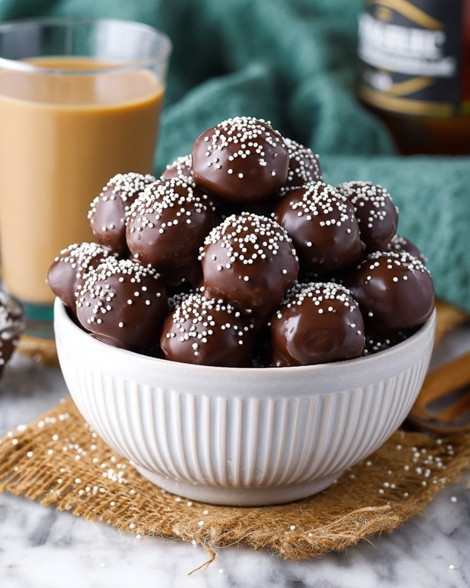 A tall pile of round chocolate balls covered with smooth, shiny dark brown chocolate and topped with small white sprinkles fills a white bowl with vertical ridges; the balls are arranged in multiple layers, with the top layer showing the most detail and texture. The bowl sits on a rustic woven mat on a white marbled surface. In the background, a glass with light brown creamy liquid is partly visible on the right side, and a blurry bottle is seen behind the bowl. Small white sprinkles are scattered on the mat and the surface around the bowl. photo taken with an iphone --ar 4:5 --v 7