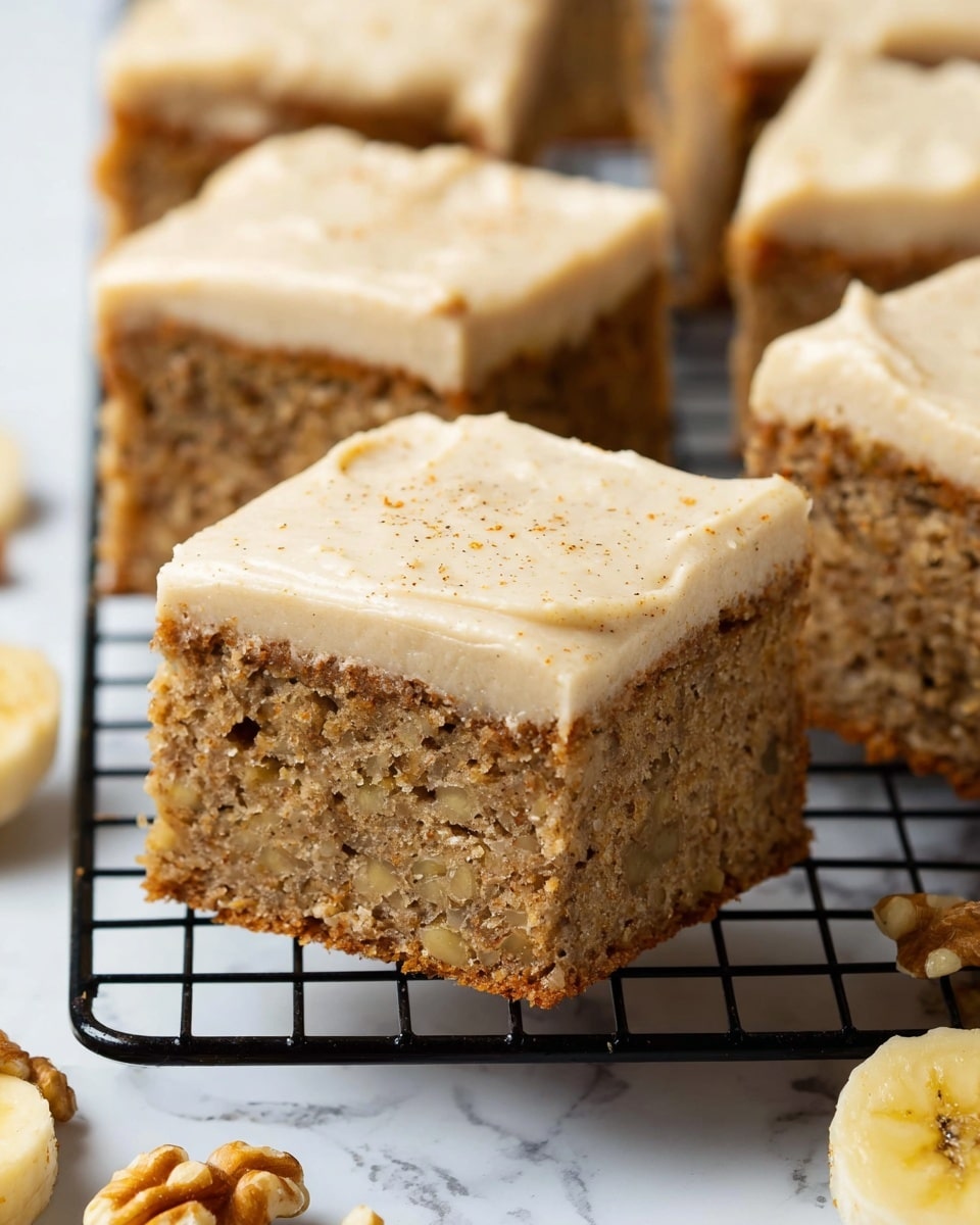 The image shows multiple square pieces of banana cake arranged closely on a black wire rack. Each piece has two layers: a thick, textured brown cake layer with visible small chunks of banana and nuts on the bottom, and a smooth, creamy light beige frosting layer on top with tiny specks of cinnamon or spice. The cake pieces sit on a white marbled surface, with some banana slices and walnut pieces scattered in the background. Photo taken with an iphone --ar 4:5 --v 7