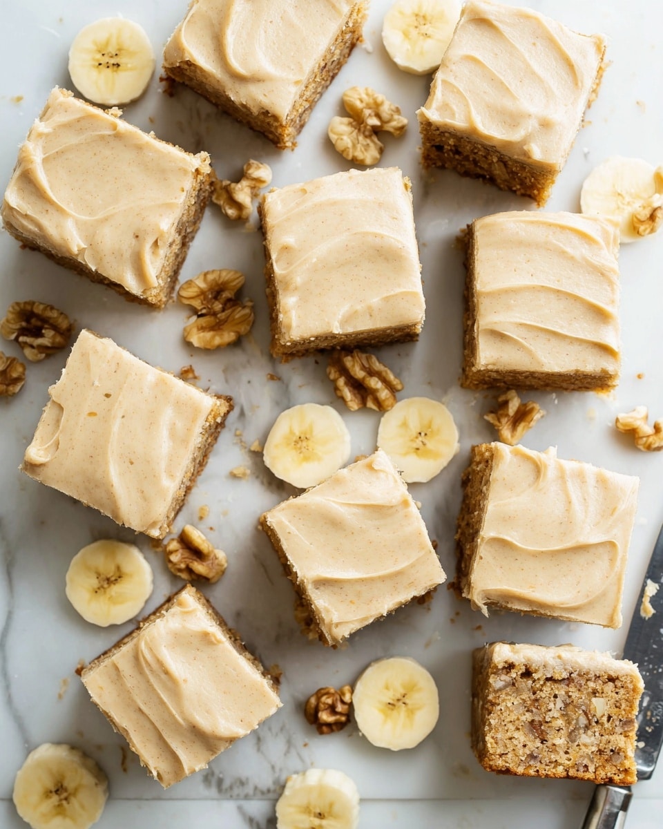 There are sixteen cut square pieces of a two-layer banana cake arranged close together on a white marbled surface. Each piece has a bottom layer that is light brown with visible small walnut chunks inside, showing a soft, moist texture, and a top layer that is a smooth, creamy beige frosting spread evenly. Around the cake pieces, there are several thin banana slices and scattered walnut halves adding a natural touch. A knife smudged with frosting lies on the right side. The light and colors make the cake look fresh and inviting. photo taken with an iphone --ar 4:5 --v 7