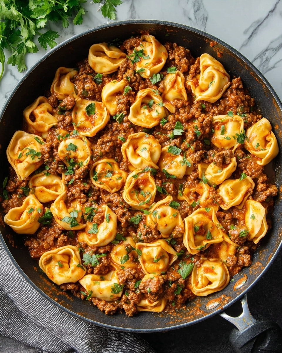 A close-up view of a black skillet filled with one layer of cooked tortellini pasta, golden yellow in color with soft, folded edges. The pasta is mixed evenly with browned ground meat pieces and covered in a reddish-orange tomato-based sauce that looks rich and slightly thick. Fresh green herb leaves, likely parsley or cilantro, are scattered on top, adding a fresh contrast to the warm colors of the dish. The skillet rests on a white marbled surface, with a gray cloth napkin to the side and some fresh herbs visible in the corner. photo taken with an iphone --ar 4:5 --v 7