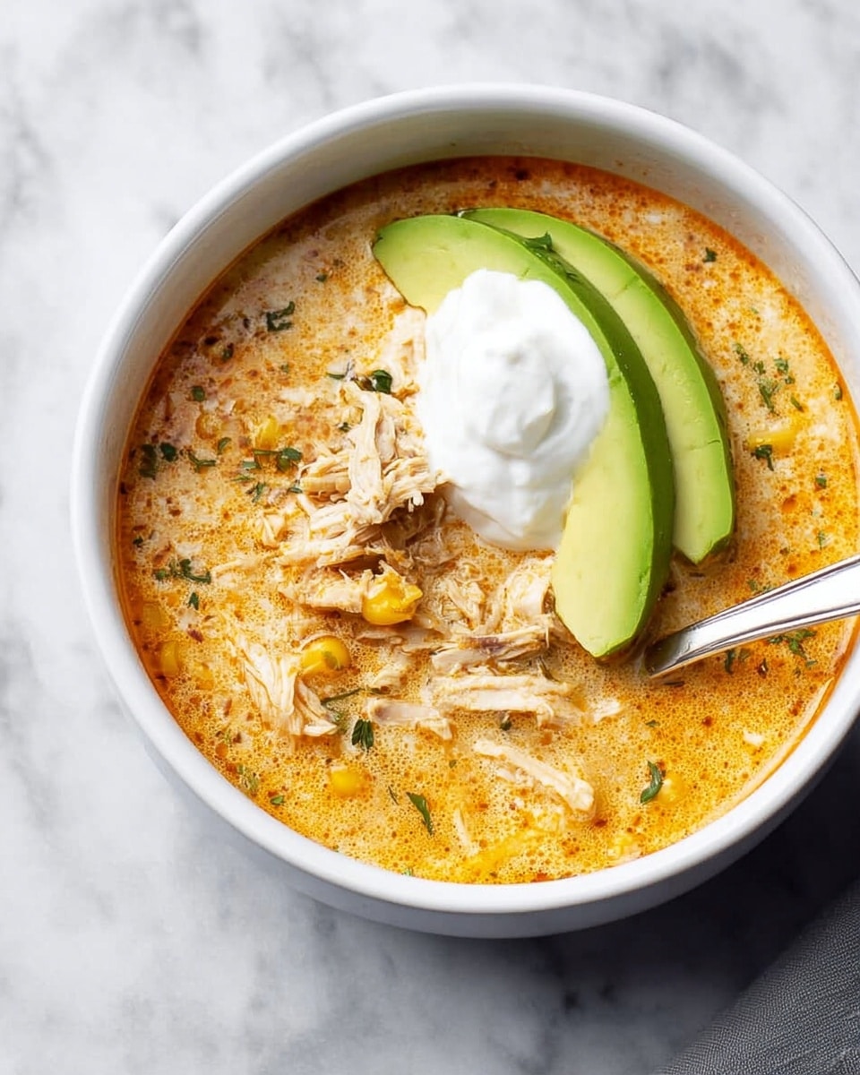 A white bowl filled with creamy soup that has a light orange color with specks of red oil on top, showing chunks of shredded chicken mixed inside. On the surface, there is a dollop of white sour cream placed near the middle, topped with a small bunch of fresh green cilantro leaves. A golden spoon rests inside the bowl on the right side. The bowl is placed on a white marbled surface with part of a white bowl in the background showing the same soup. A white cloth is visible in the lower left corner of the image. photo taken with an iphone --ar 4:5 --v 7