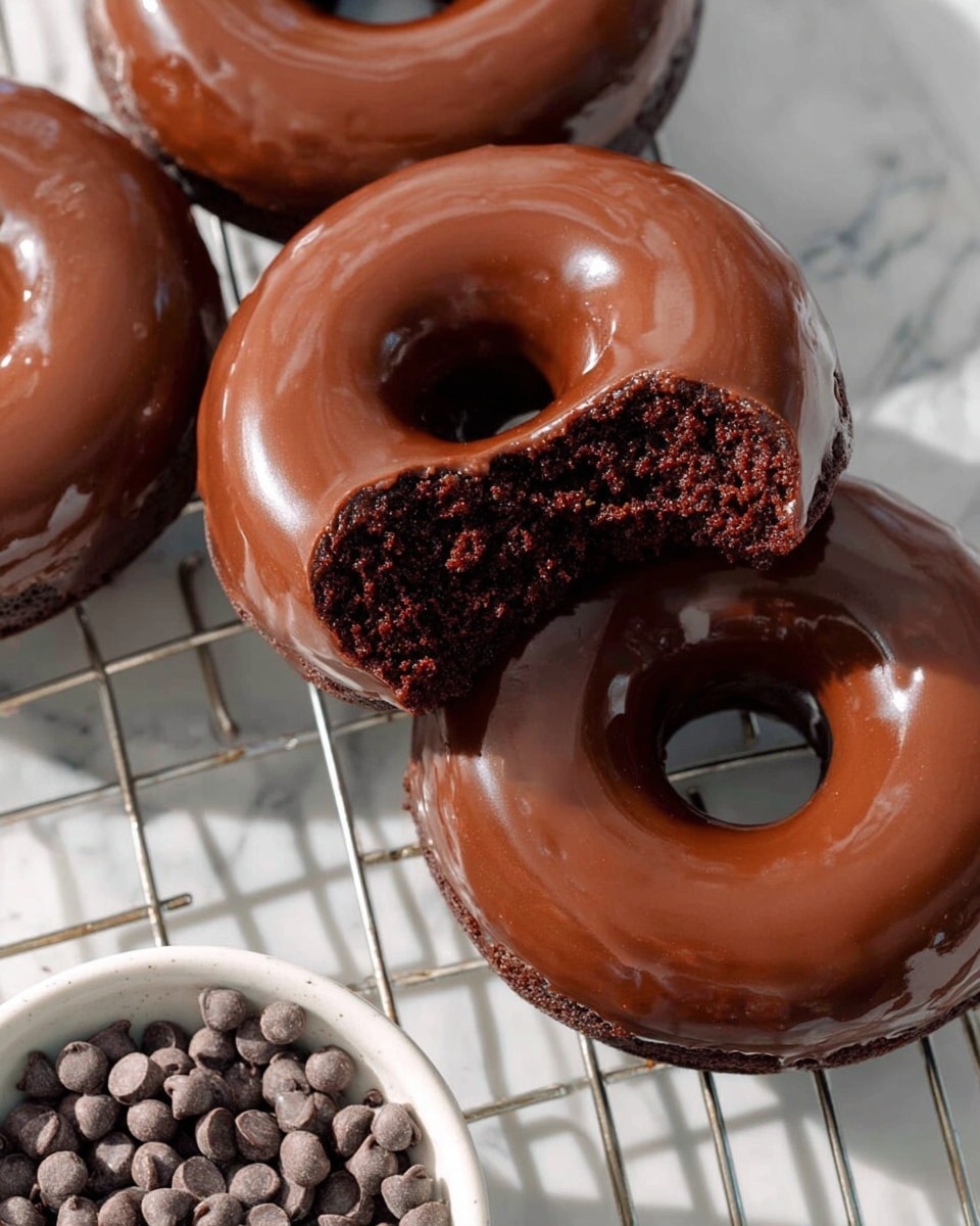 The image shows three chocolate donuts on a cooling rack with shiny, smooth chocolate glaze covering the whole top surface of each donut. One donut is in the center with a bite taken out, showing a dark, moist, and crumbly chocolate interior with a soft texture. The donuts have a rich dark brown color under the glaze, and the glaze catches light, creating a glossy look. A white bowl filled with small chocolate chips is placed on a white marbled surface near the bottom left corner of the image. The scene is bright, with natural light highlighting the donuts' texture and shine. photo taken with an iphone --ar 4:5 --v 7