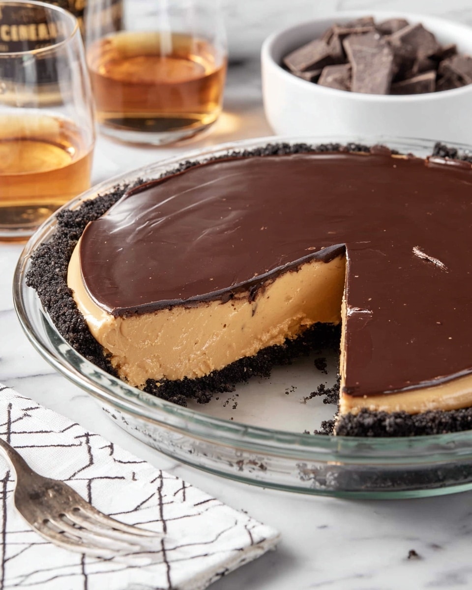 A round pie in a clear glass dish with three layers: the bottom crust is dark crumbly chocolate, the middle layer is thick and smooth light tan peanut butter filling, and the top is a shiny, dark chocolate ganache spread evenly. One slice is cut but still in place, showing the clean edges of each layer. The dish sits on a white marbled surface with a white and black geometric napkin and a fork nearby. In the background, there is a glass of amber liquid and a white bowl filled with small dark chocolate pieces. Photo taken with an iphone --ar 4:5 --v 7