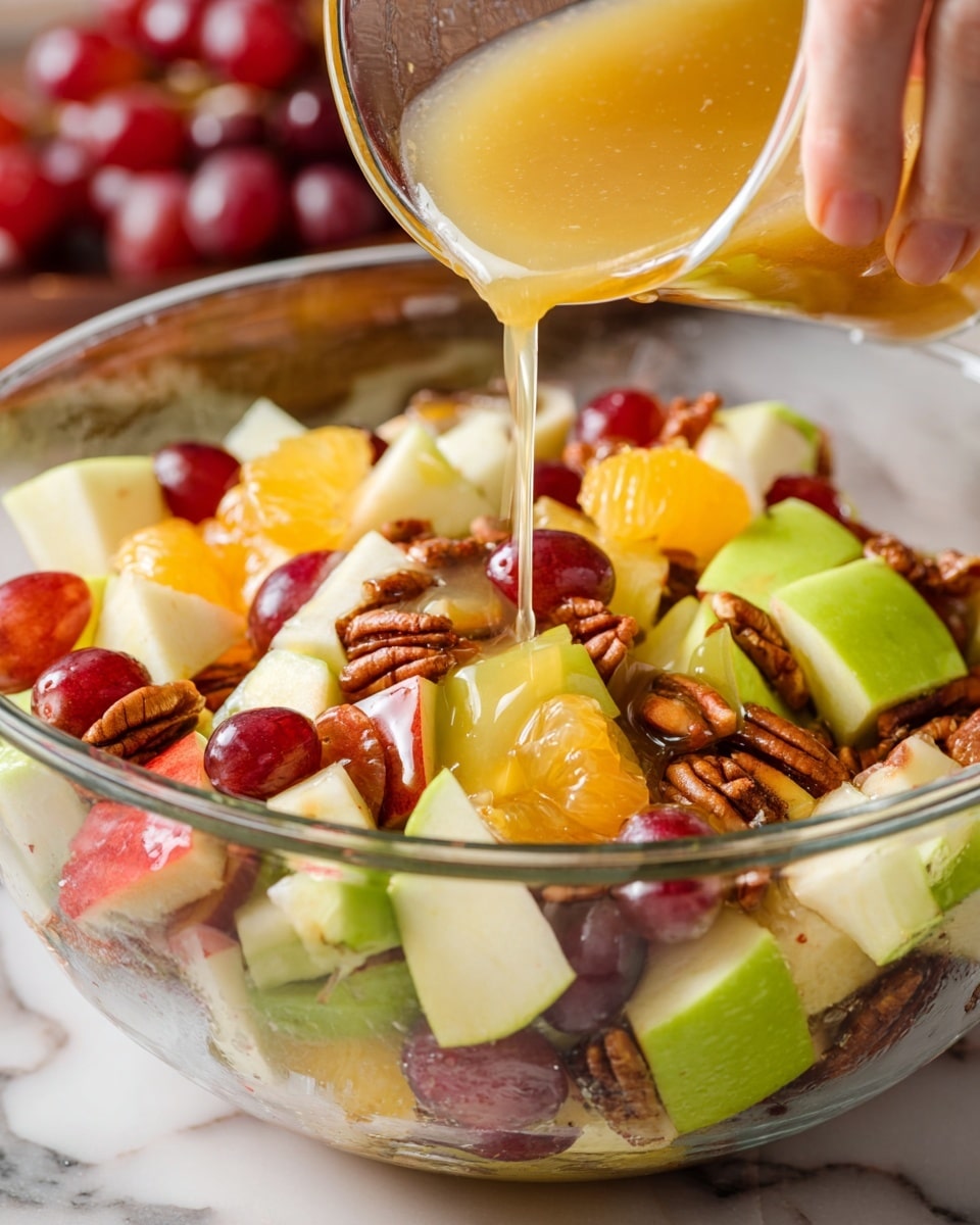 A clear glass bowl filled with a colorful fruit salad sits on a white marbled surface. The salad has many layers of small fruit pieces: green apple chunks with smooth skin, red apple pieces with a light shine, bright orange segments, and red grapes. Mixed in are brown pecan nuts and small bits of dark red dried cranberries, adding texture. Around the bowl are a whole green apple, a sliced green apple showing its light inside, scattered pecans, and a small clear bowl of red grapes. A green and white checkered cloth and wooden salad utensils are nearby, all on a white marbled surface. Photo taken with an iphone --ar 4:5 --v 7