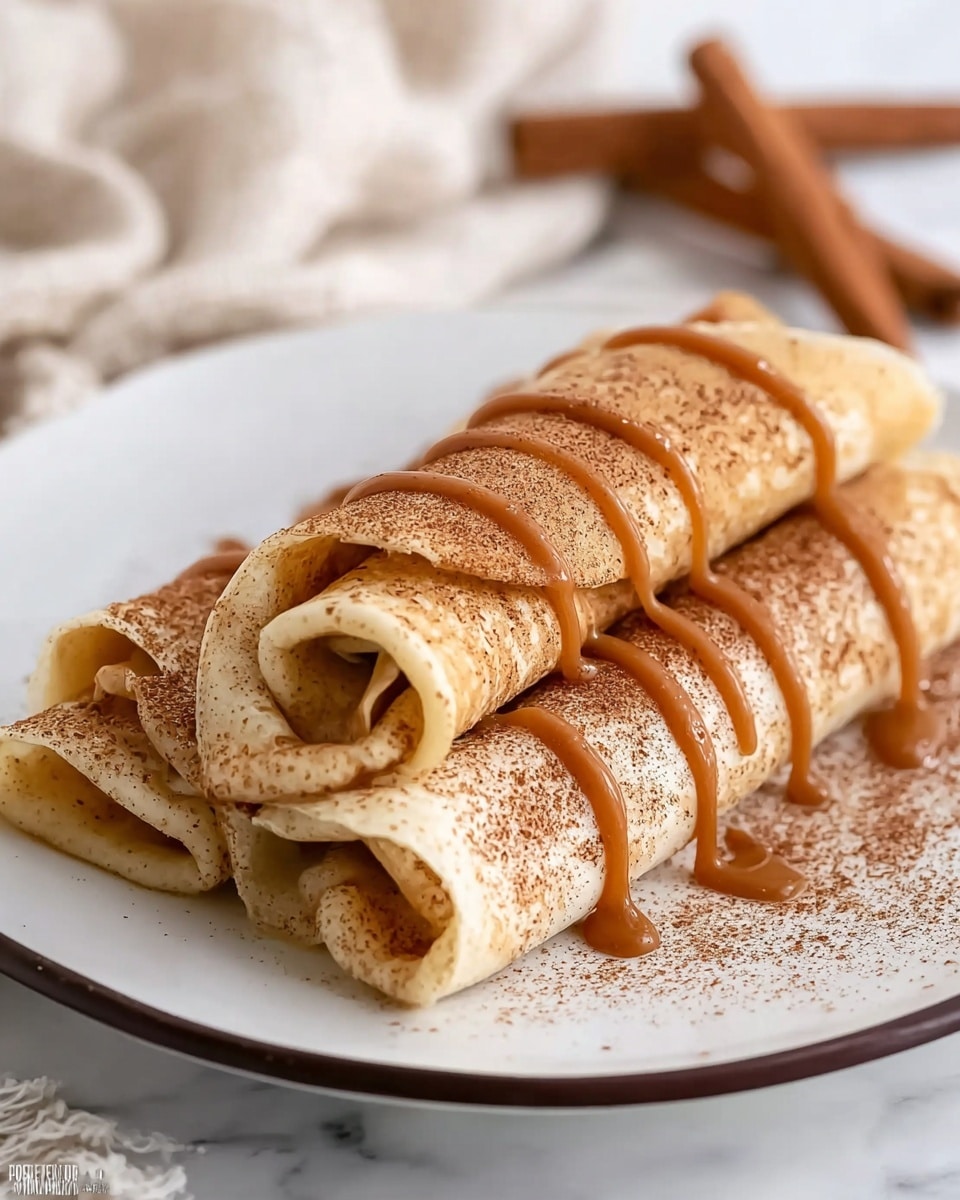 The image shows four rolled crepes stacked in a pyramid shape on a white plate with a thin black rim, placed on a white marbled surface. Each crepe is light golden in color with a soft texture and sprinkled evenly with brown cinnamon powder. Drizzled over the top crepes is a thick, smooth light brown caramel sauce that flows in ribbons down the sides. The crepes are tightly rolled, revealing a small hollow center in each. The background is blurred white fabric, enhancing the focus on the food. photo taken with an iphone --ar 4:5 --v 7