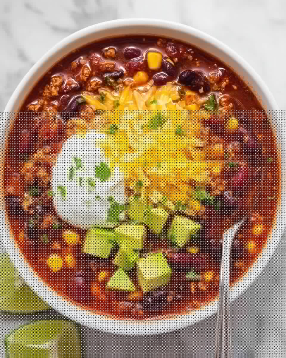 A white bowl filled with a thick chili made of dark red kidney beans, black beans, yellow corn, and small pieces of beige ground meat mixed in a rich red tomato sauce. On top, bright yellow shredded cheddar cheese is piled over a dollop of smooth white sour cream, surrounded by green cube-shaped avocado chunks and sprinkled with small green cilantro leaves. A fresh green lime wedge rests partly submerged on the left side of the bowl, and a silver spoon is placed on the right side, touching the chili. The bowl is set on a white marbled surface. photo taken with an iphone --ar 4:5 --v 7