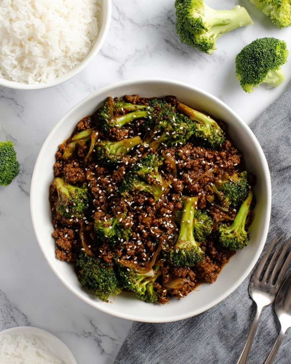 The image shows a white bowl filled with a dish made of cooked ground meat mixed with bright green broccoli florets, all coated in a thick, dark brown sauce with visible pieces of cooked onions throughout. On top, there is a light sprinkling of sesame seeds adding texture. To the top left, there is a white bowl with fluffy white rice, and the background is a white marbled surface with loose broccoli stems and a grey cloth underneath the dishes. At the bottom right corner, there is a white plate with a spoon and fork resting on it. Photo taken with an iphone --ar 4:5 --v 7