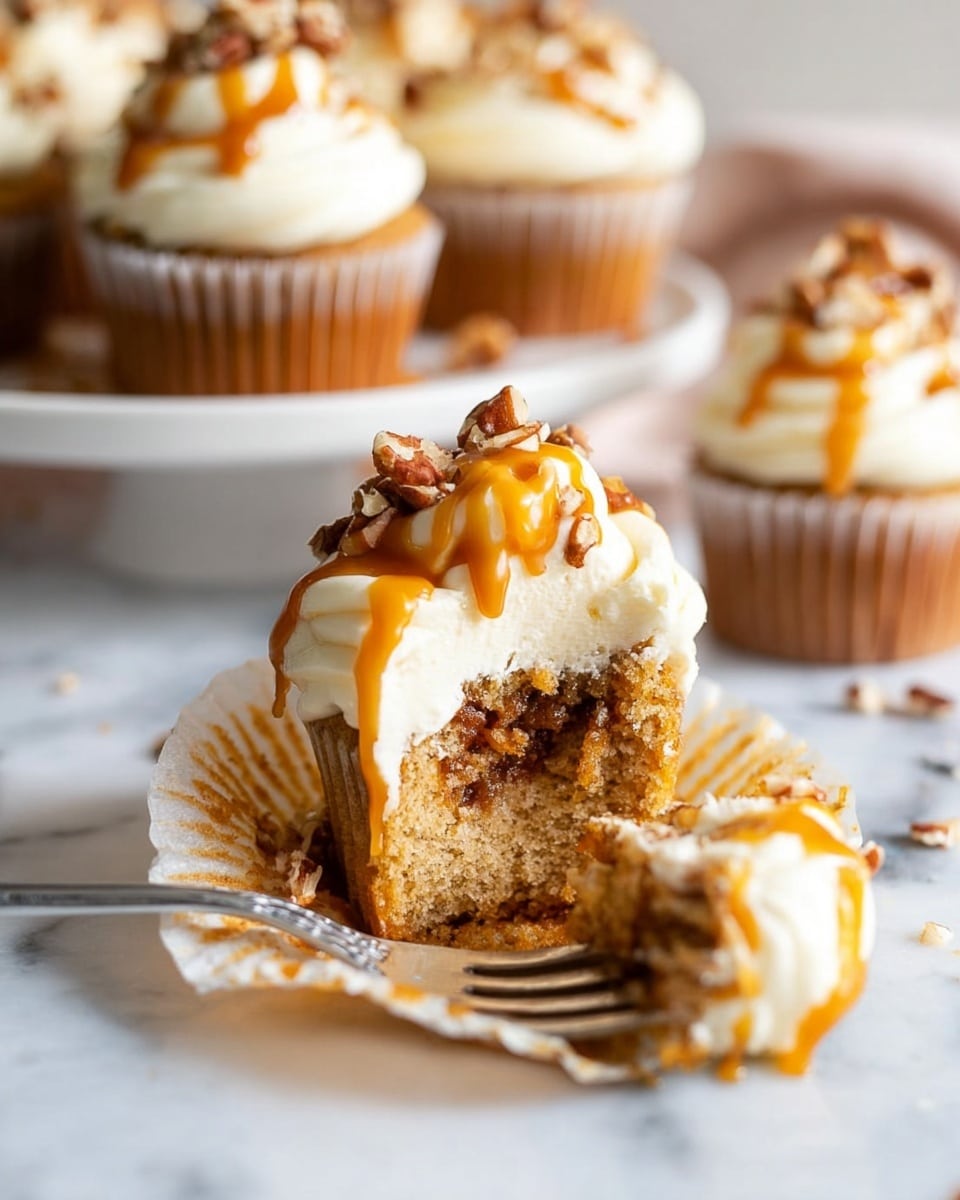A single cupcake standing on a white marbled surface, with three visible layers: the base is a brown textured cake wrapped in a white ridged paper liner, the middle layer is a thick, smooth white frosting domed on top of the cake, and the top layer is a drizzle of shiny golden caramel sauce flowing over the frosting and slightly down the sides, sprinkled with small chunks of crumbly brown nuts or streusel. The background is softly blurred with warm light, showing more similar cupcakes out of focus. photo taken with an iphone --ar 4:5 --v 7