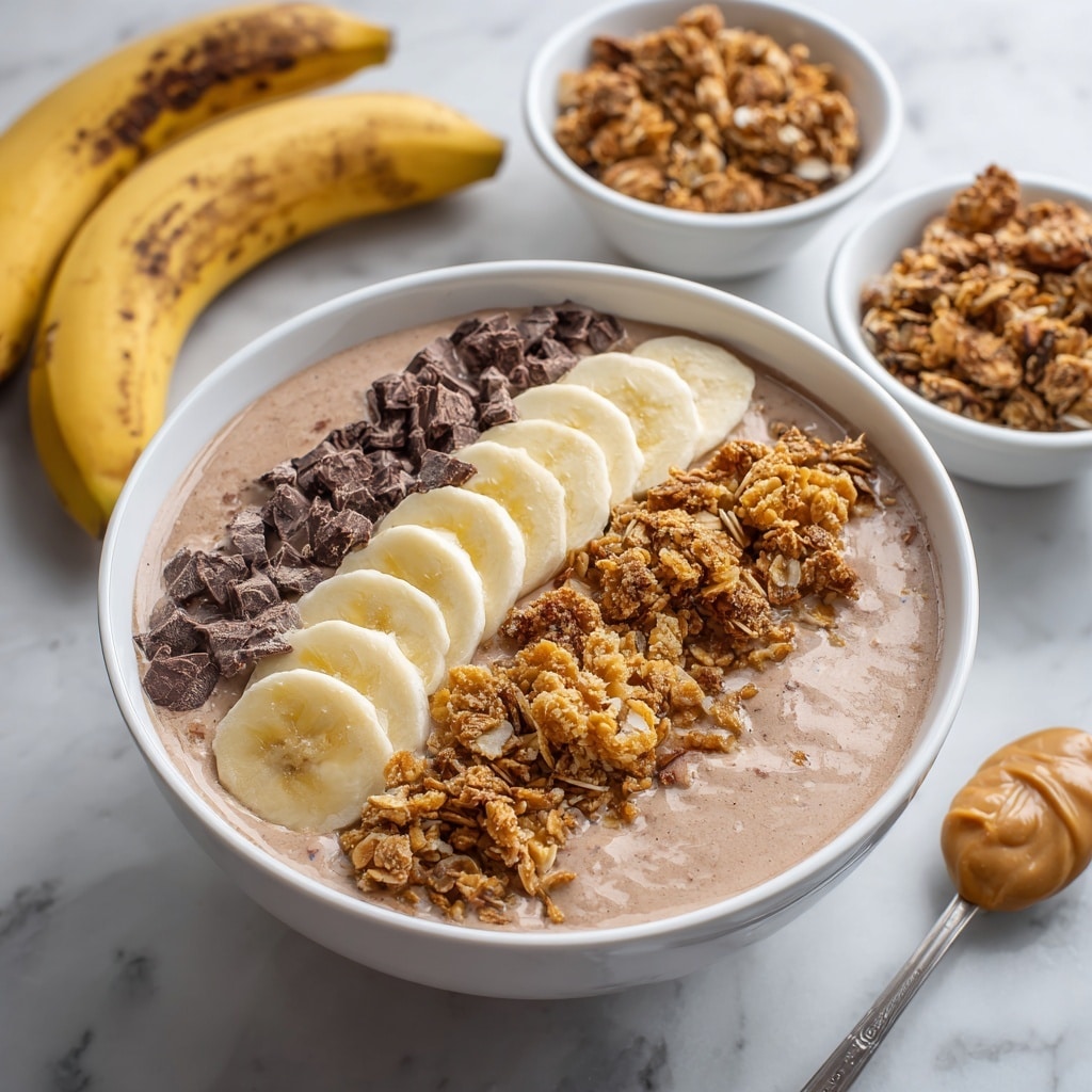 A white bowl filled with a smooth light brown smoothie base, topped with three neat horizontal layers on the top: dark brown chopped chocolate pieces at the far left, pale yellow banana slices in the middle, and golden brown crunchy granola on the right. The bowl sits on a white marbled surface with scattered chocolate chunks nearby and a spoon with creamy peanut butter resting in front of the bowl. In the background, a white bowl with granola pieces and a whole banana are softly blurred. Photo taken with an iphone --ar 4:5 --v 7