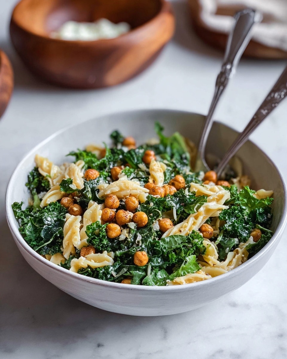 A white shallow bowl contains a fresh pasta salad with three main layers: at the bottom are dark green curly kale leaves with a rough texture, scattered evenly; the middle layer shows twisted short pasta pieces, pale yellow and smooth, resting on the kale; on top there are small round crispy golden-brown chickpeas spread across the dish. Two metal forks stand in the bowl among the salad. The bowl is placed on a white marbled surface, with a blurred wooden bowl in the background holding a white creamy substance. Photo taken with an iphone --ar 4:5 --v 7