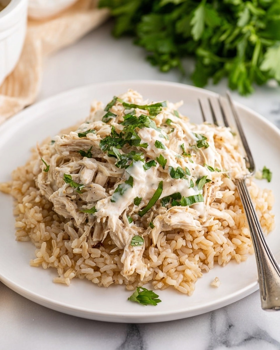 A white plate holds a base layer of cooked rice, light brown and fluffy, covering the entire plate. On top, there is a generous layer of shredded chicken mixed with a creamy sauce that has a pale beige color with small bits of herbs and onions visible. The chicken mixture looks moist and tender with some green parsley pieces sprinkled throughout. A pair of metal tongs lifts some of the chicken mixture, showing its soft texture. The plate is set on a white marbled surface, and in the background, there is a blurred white slow cooker with a dial. Photo taken with an iphone --ar 4:5 --v 7