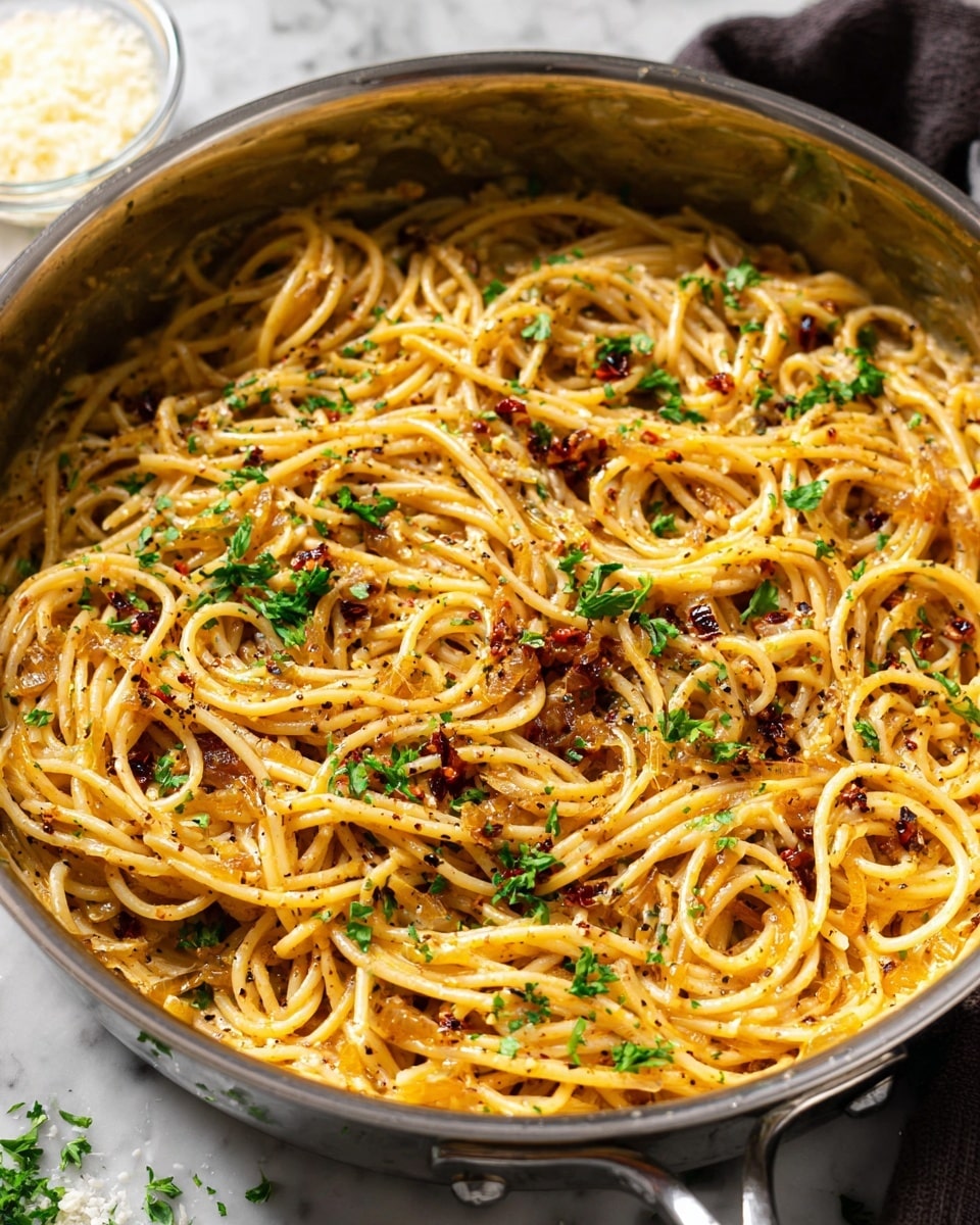 A close-up view of a shiny stainless steel pan filled with creamy spaghetti pasta. The pasta is light golden yellow with a smooth, slightly oily texture and is mixed well with caramelized onion bits and small flecks of dark red chili flakes. Freshly chopped green parsley pieces are scattered evenly on top, adding a fresh, vibrant touch. The sauce coating the pasta gives it a glossy, rich look with some black pepper specks visible throughout. In the background, there is a small glass bowl with grated Parmesan cheese on a white marbled surface. photo taken with an iphone --ar 4:5 --v 7