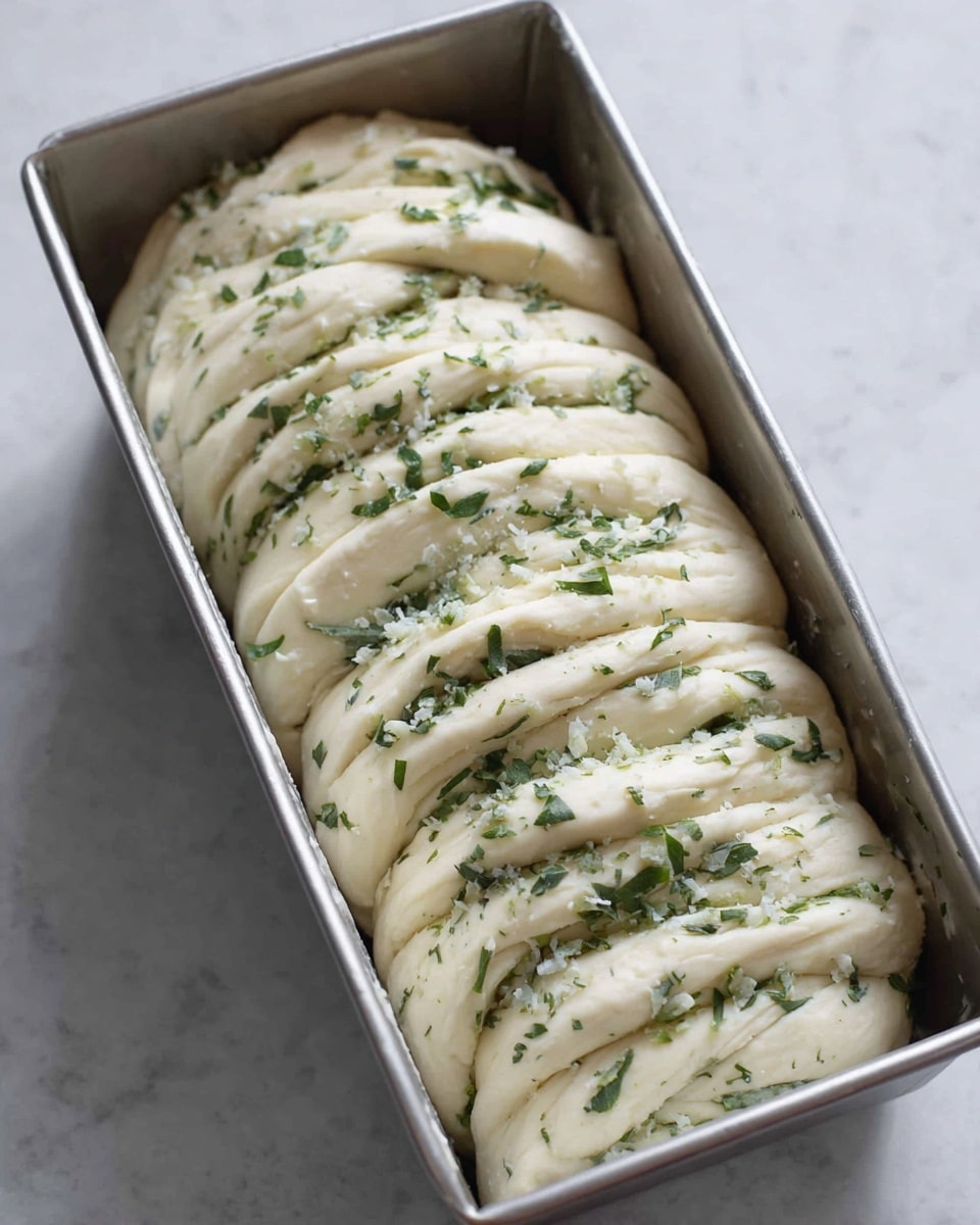 This image shows a metal loaf pan filled with many layers of soft dough folded closely together. Each dough layer is pale and smooth with small bits of green herbs and white flakes sprinkled on and between the layers, creating a textured look. The dough looks fluffy and fresh, with the herbs adding a vibrant pop of green throughout. The pan sits on a white marbled surface, enhancing the clean, fresh feel of the image. photo taken with an iphone --ar 4:5 --v 7