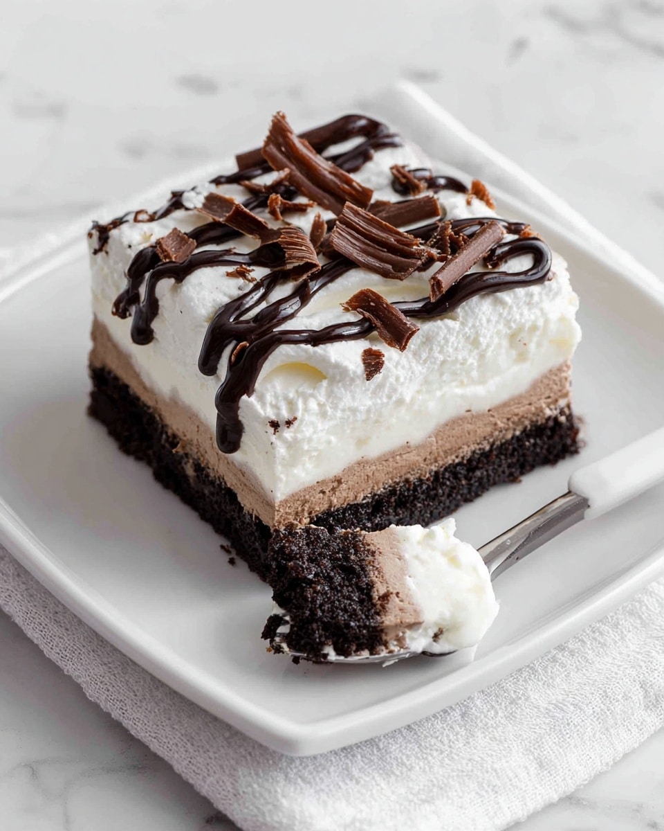 A square layered dessert is placed on a white plate on a white marbled texture. The dessert has three clear layers: a bottom dark brown chocolate cake layer, a smooth middle light brown cream layer, and a top fluffy white cream layer. The top white cream is decorated with dark chocolate drizzle and small chocolate shavings scattered over it. Extra chocolate drizzle is artistically spread on the plate around the dessert. The background shows a white baking dish with more of the same dessert cut into squares, visible in the upper left corner. Photo taken with an iphone --ar 4:5 --v 7