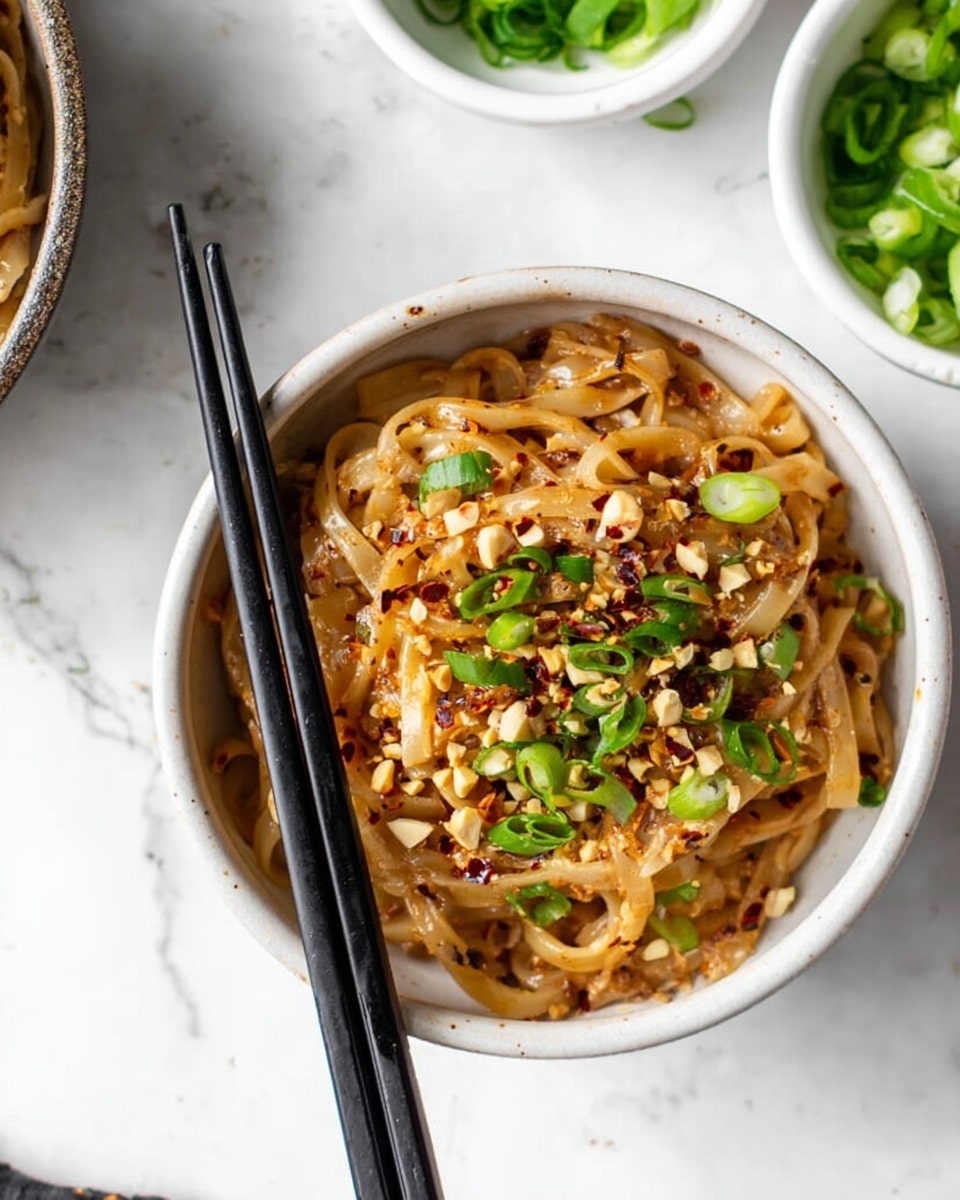 A white bowl filled with thick noodles coated in a light brown sauce, topped with sliced green onions and crushed peanuts scattered across the surface, with small red chili flakes sprinkled over everything. A pair of black chopsticks rests inside the bowl on the left side. The bowl sits on a white marbled surface with parts of another bowl and a small white bowl of chopped green onions visible in the background. photo taken with an iphone --ar 4:5 --v 7