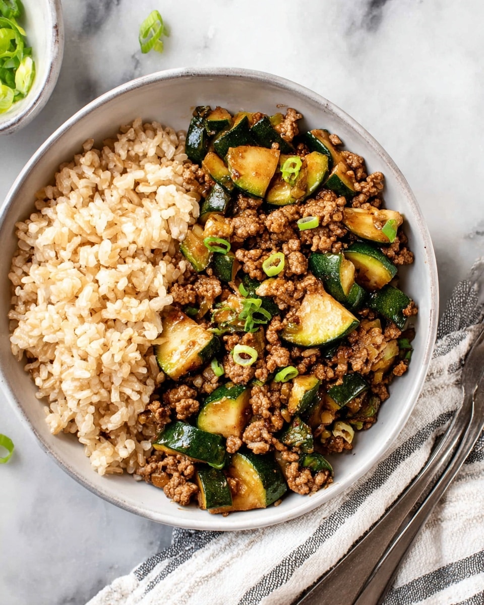 A round white bowl holds two layers of food placed side by side. The left layer is light brown cooked rice with a soft, fluffy texture. The right layer consists of cooked ground meat mixed with chopped green zucchini pieces; the meat is brown and crumbly, while the zucchini is green with a glossy, cooked surface. Small bits of green onions are scattered on top. The bowl is set on a white marbled surface, with a silver fork and a white cloth with gray stripes on the lower right side. Photo taken with an iphone --ar 4:5 --v 7