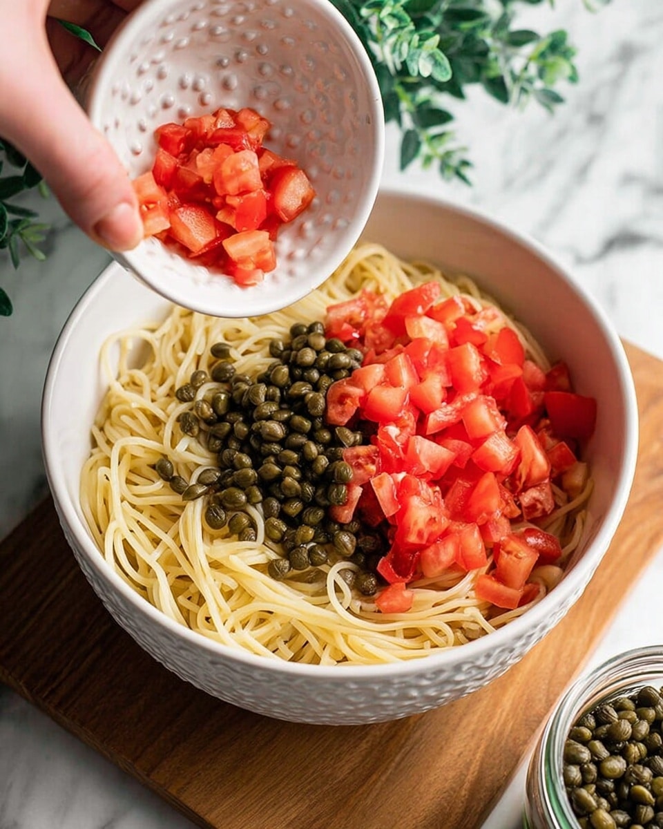 A white bowl filled with spaghetti pasta as the base layer, light yellow and smooth in texture, topped with small red tomato chunks scattered evenly. There are green capers and parsley leaves sprinkled on top, adding different shades of green and texture contrast. Thin yellow lemon zest strips are evenly spread across the surface. The bowl is set on a white marbled surface with a soft green cloth nearby and two whole and half-cut lemons blurred in the background. photo taken with an iphone --ar 4:5 --v 7