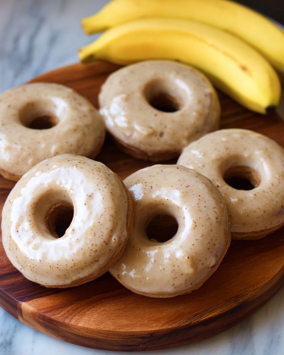 A woman's hand is holding a round donut covered with smooth, light beige icing that has tiny dark specks, creating a creamy texture on top. The donut’s base is light brown with a soft, fluffy inside. Behind the donut, there are two more donuts, one close and one a bit farther, both plain without icing. Below the held donut, a white bowl filled with the same beige icing, slightly shiny and thick, is visible. All items rest on a white marbled surface that adds a clean, bright background to the warm, soft colors of the donuts and icing. Photo taken with an iphone --ar 4:5 --v 7