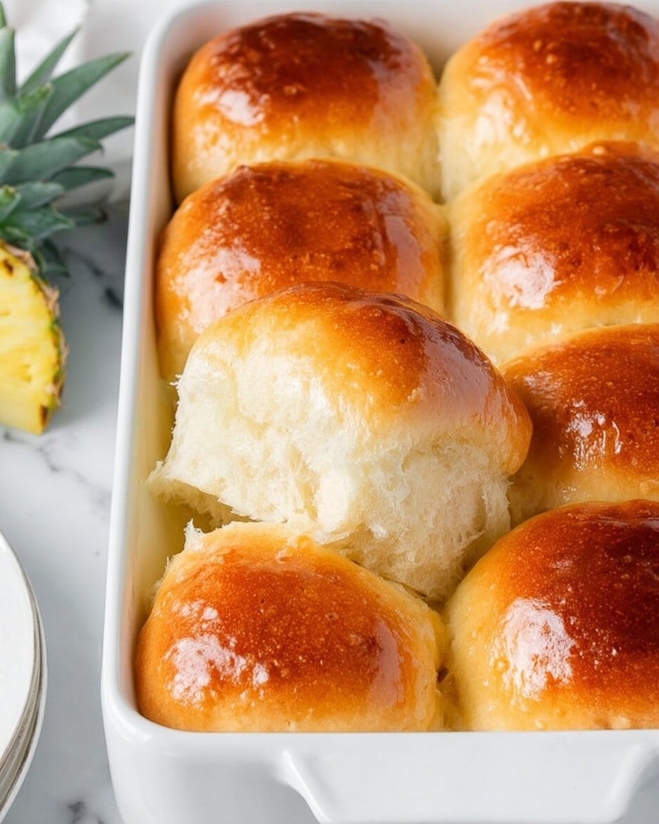 The image shows a white rectangular baking dish containing six soft, golden brown dinner rolls closely packed together. The rolls have a shiny, smooth top crust with a rich golden color, slightly darker in some places due to baking. One roll is pulled away, revealing a fluffy and light interior with a soft, airy texture. In the background, part of a pineapple crown can be seen on a white marbled surface, with a corner of a white plate also visible. The overall scene is bright and clean with a fresh and inviting look. photo taken with an iphone --ar 4:5 --v 7