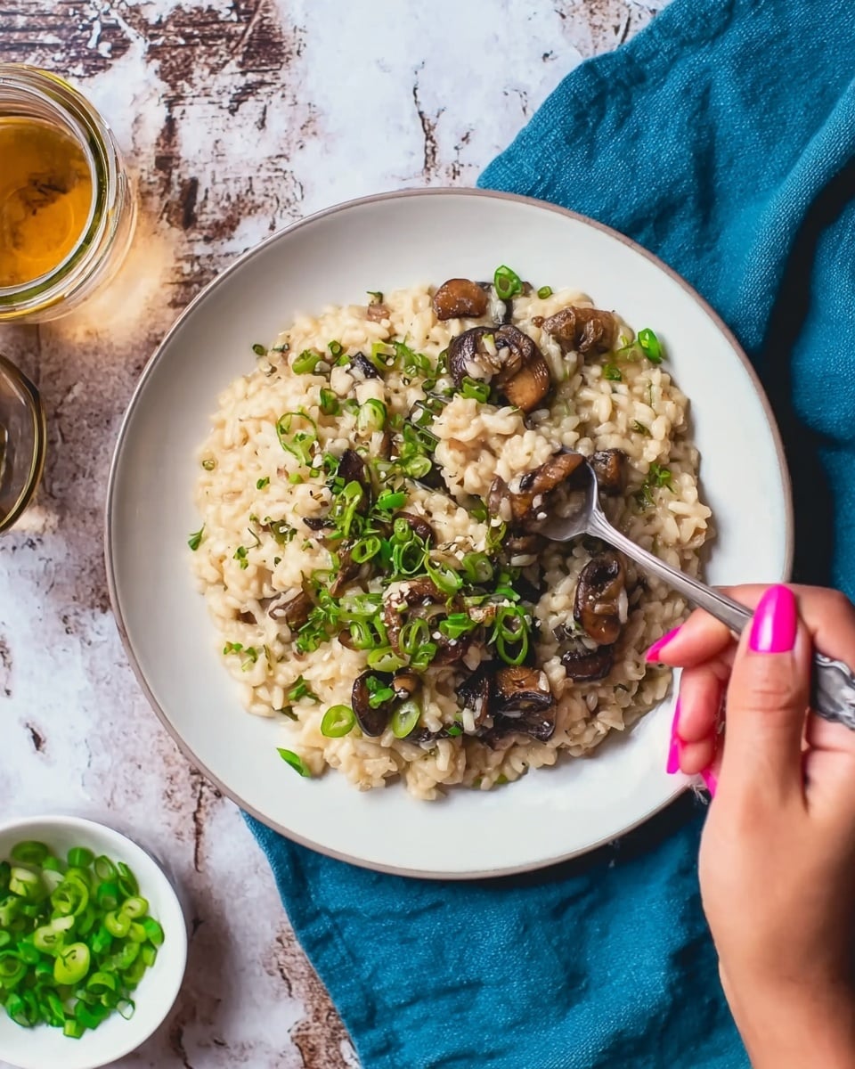 A close-up view of a creamy risotto served in a white plate with a rustic brown rim, resting on a wooden board over a white marbled texture with a blue cloth underneath. The risotto has a soft, creamy texture with visible grains of rice mixed with browned mushrooms scattered throughout. The dish is topped with finely chopped bright green onions and delicate microgreens, adding a fresh touch. A silver spoon is positioned inside the plate on the right side, partially submerged in the risotto, enhancing the inviting appearance. In the background, there is a small white bowl filled with chopped green onions and a wooden salt container. Photo taken with an iphone --ar 4:5 --v 7