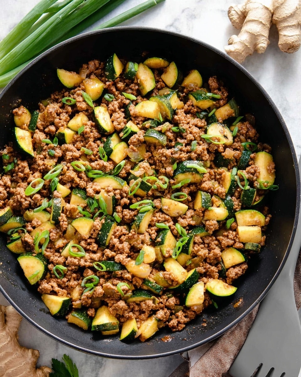 A close-up view of a black skillet filled with cooked ground meat mixed with small pieces of sautéed zucchini and onions, all evenly cooked to a light brown and golden color, garnished with sliced green onions scattered on top. The zucchini pieces are cut into half-moon shapes, showing a green outer skin and pale inside. The skillet is set on a white marbled surface with fresh green onions and ginger root nearby, along with a gray spatula resting beside the skillet. photo taken with an iphone --ar 4:5 --v 7
