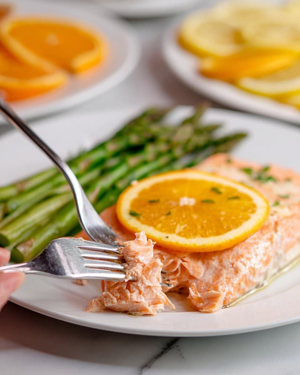 A close-up image shows a white plate with a fillet of cooked salmon topped with a bright orange slice in the center. Behind the salmon, a small bunch of green asparagus spears is placed neatly. A woman's hand holding a fork lifts a piece of the pink, flaky salmon close to the camera. The plate is set on a white marbled surface, and in the blurry background, another white plate holds slices of orange and lemon. photo taken with an iphone --ar 4:5 --v 7