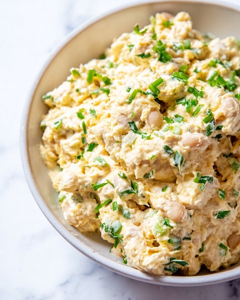 A sandwich cut in half and stacked, showing two thick layers of creamy tuna salad mixed with small green bits and topped with fresh bright green sprouts. Each half is between two slices of light brown whole grain bread with a soft, coarse texture. The sandwich sits on a light wooden board with scattered green sprouts around. The background has a white marbled texture with a white bowl filled with more tuna salad slightly out of focus. Photo taken with an iphone --ar 4:5 --v 7