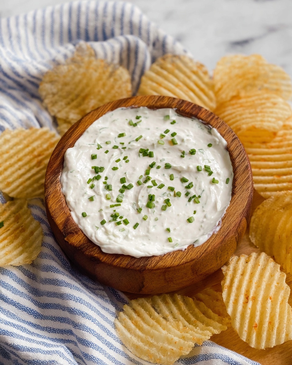 A woman's hand is dipping a ridged light golden potato chip into a small wooden bowl filled with thick white dip speckled with small green chive pieces, some of which float on top of the dip. The bowl is placed on a white marbled surface with a blue and white woven cloth beneath it. More potato chips are scattered around the bowl, showing their ridged texture and pale yellow color. Photo taken with an iphone --ar 4:5 --v 7