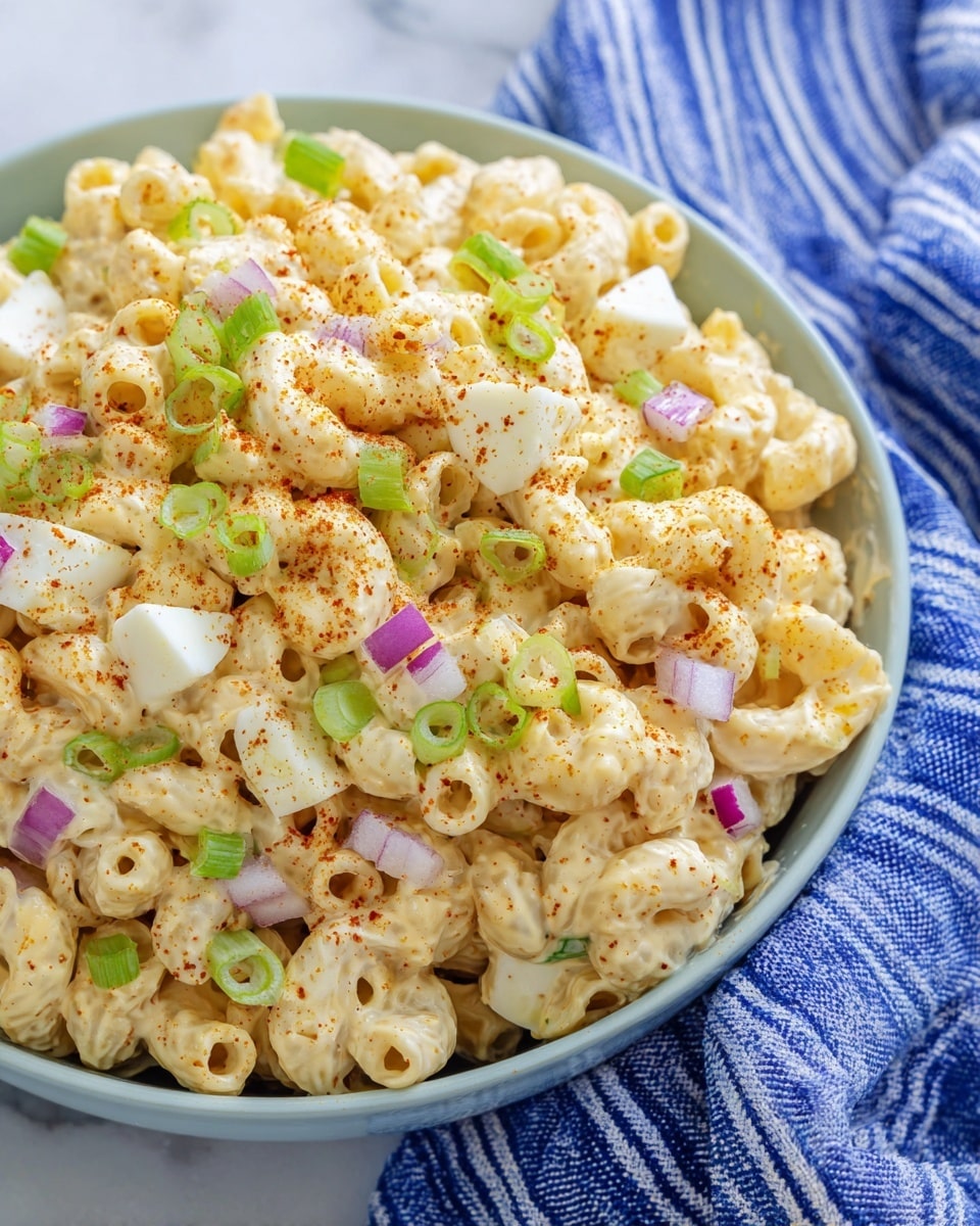 A close-up view of a creamy pasta salad served in a white bowl, filled with three layers of small, tube-shaped macaroni pasta coated in a smooth, light yellow dressing. Scattered on top are small pieces of white boiled egg, finely chopped purple onion, and thin slices of bright green scallions, adding pops of color. The dish is sprinkled lightly with red paprika powder for a touch of spice. The bowl is placed on a white marbled surface with a blue and white striped cloth partially visible in the background. photo taken with an iphone --ar 4:5 --v 7
