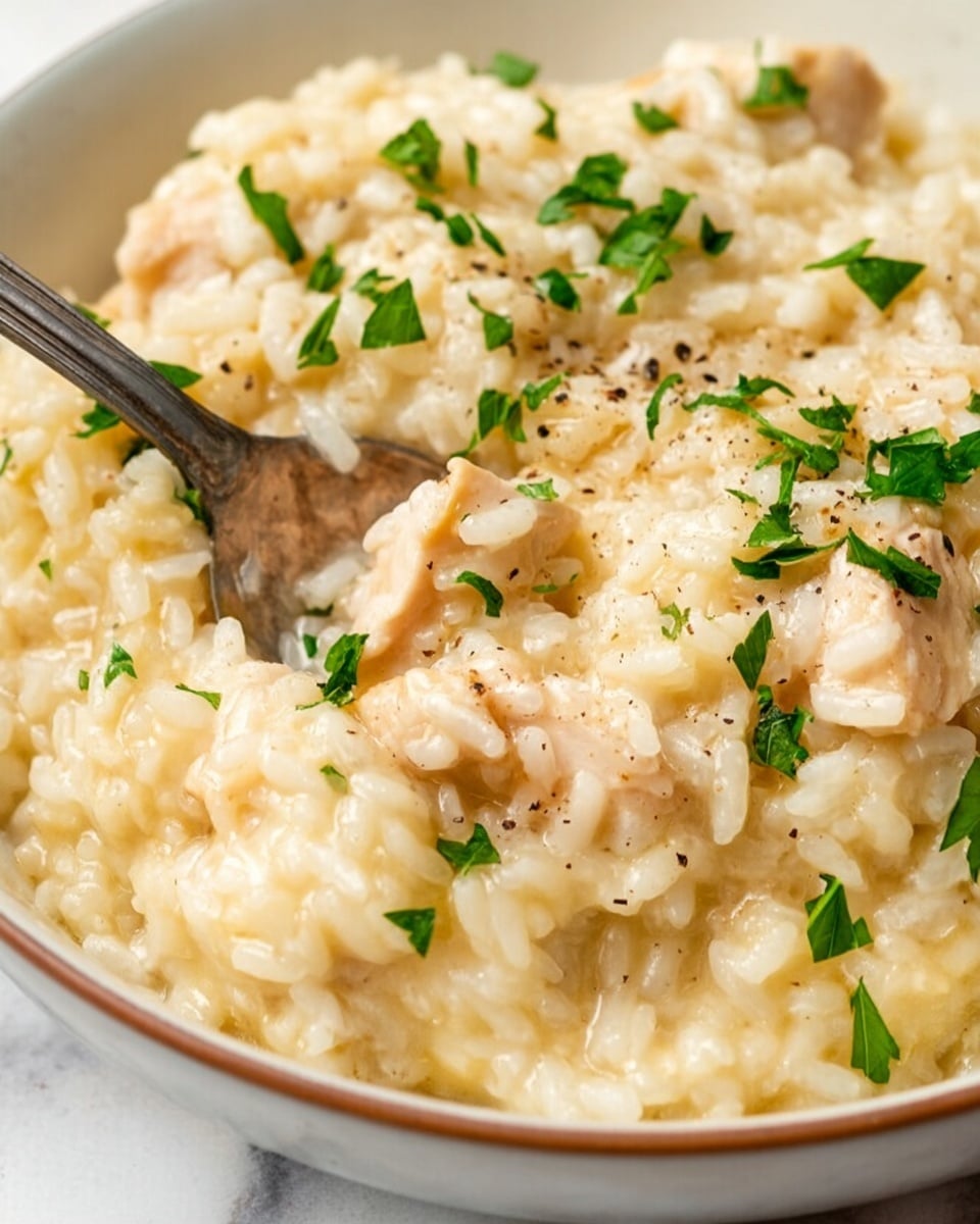 A close-up view of creamy risotto served in a white bowl, showing soft, slightly glossy cooked rice grains mixed with tender, light beige pieces of chicken. The dish is sprinkled with small, bright green chopped parsley and a few coarse cracks of black pepper scattered evenly on top. A silver spoon rests inside the bowl, partly covered by the risotto. The background is a white marbled texture. photo taken with an iphone --ar 4:5 --v 7