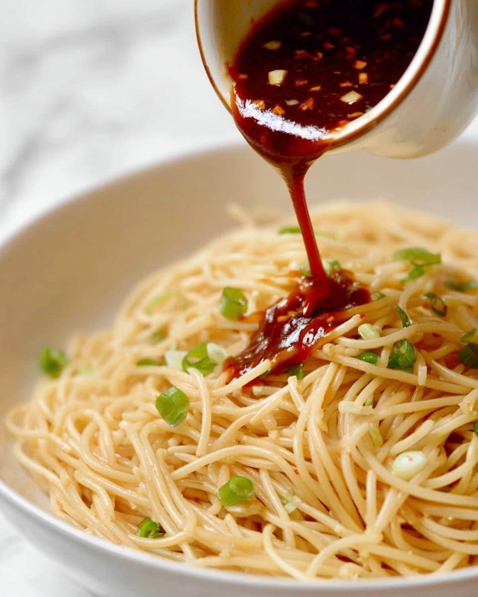 A close-up of a white bowl filled with plain cooked spaghetti noodles mixed with small pieces of chopped green onions scattered evenly on top. A small white pouring vessel with a brown rim is tilted above the noodles, with a dark red sauce flowing out in a thin stream onto the spaghetti. The sauce has a thick texture with visible small chunks inside. The background shows a white marbled surface. photo taken with an iphone --ar 4:5 --v 7