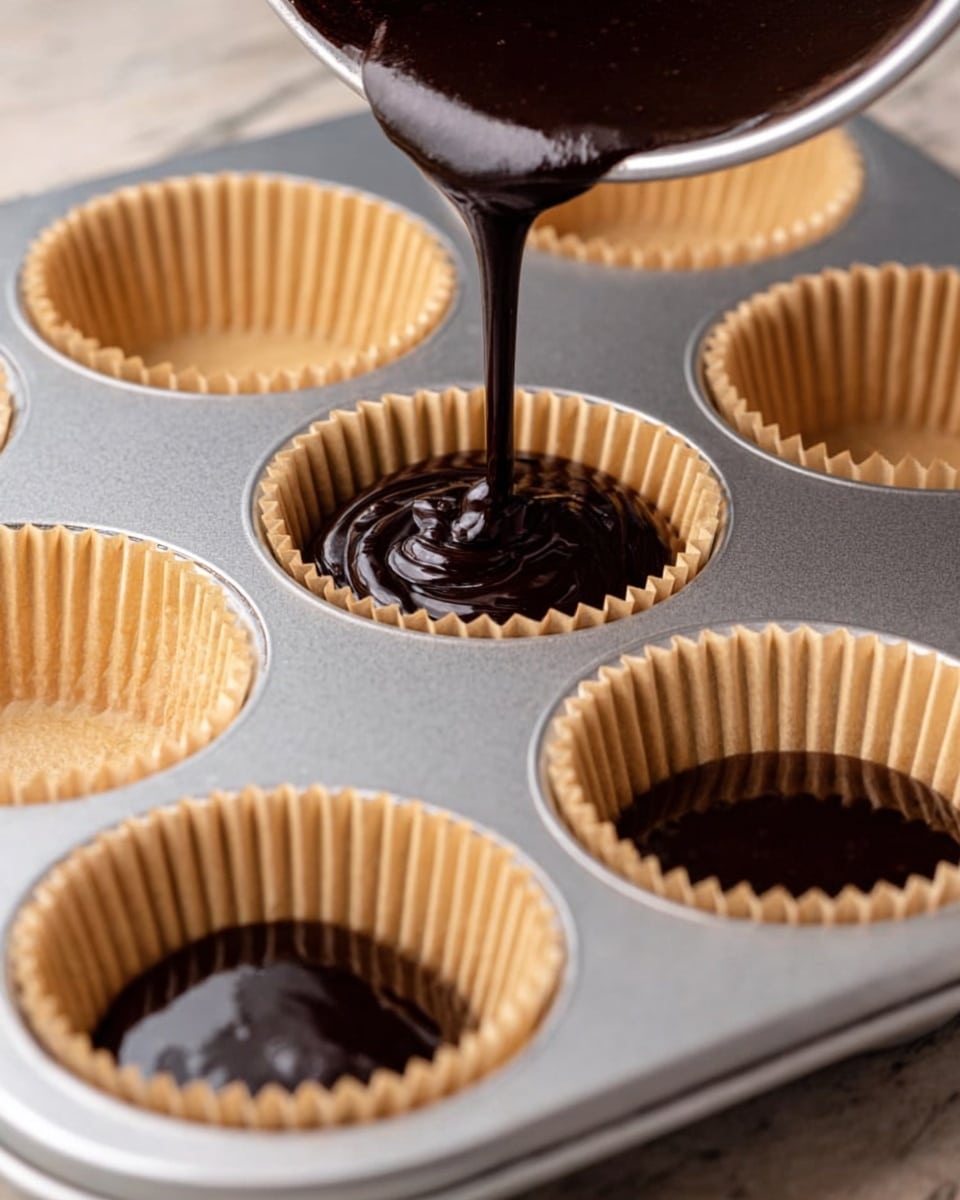 The image shows a close-up of a dark chocolate cupcake with rich chocolate frosting. The cupcake has two layers: the bottom is a moist, dark brown cake layer with a crumbly texture, and the top is a thick, smooth, and creamy chocolate frosting layer in a slightly lighter brown shade. The frosting is sprinkled with small purple and white sugar crystals around the edges. The cupcake liner is a natural light brown color and peeled away slightly. The background is out of focus with more cupcakes and a dark textured surface beneath, changed to a white marbled texture. Photo taken with an iphone --ar 4:5 --v 7