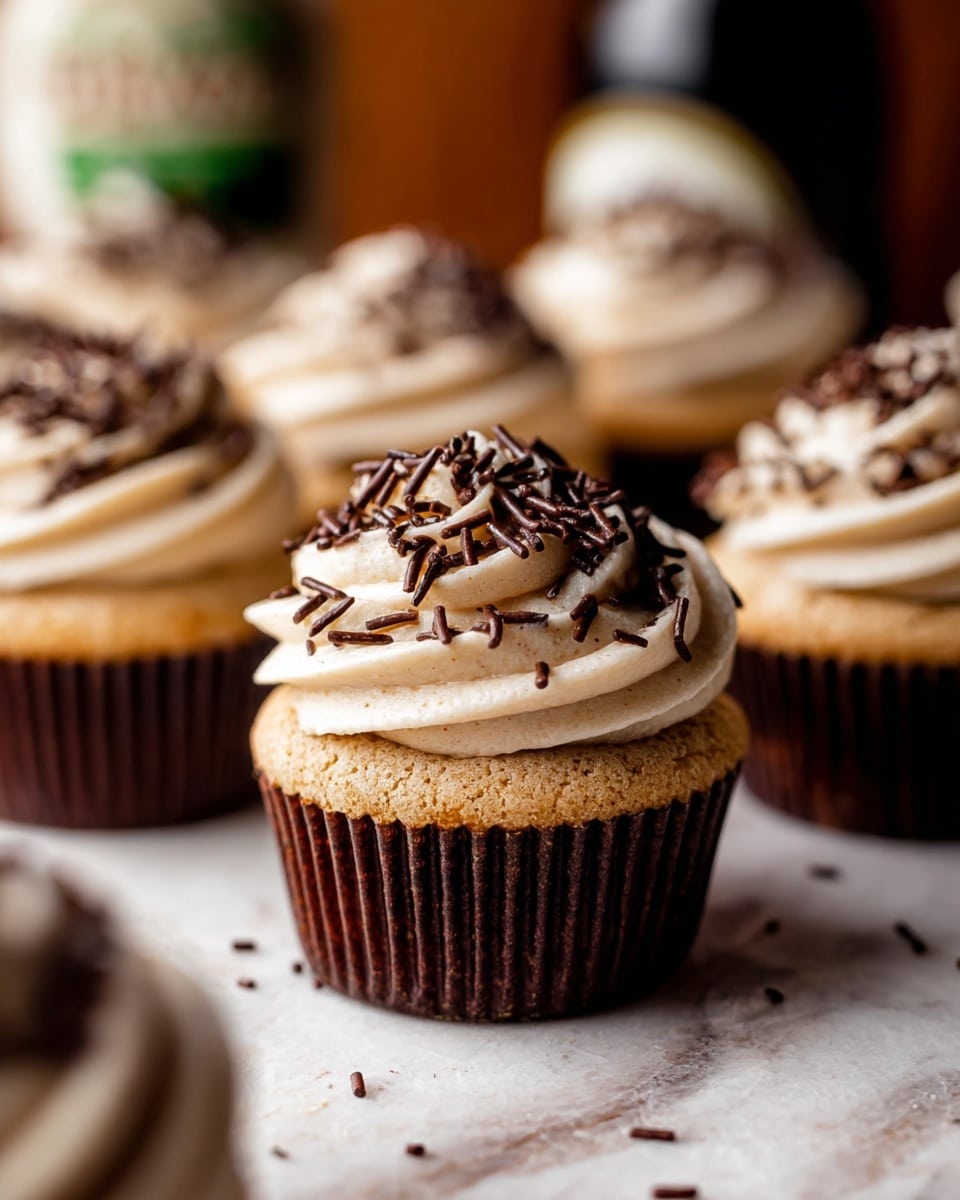 The image shows close-up of several cupcakes arranged on a white marbled surface. Each cupcake has one layer of golden brown cake at the base wrapped in a dark brown ridged paper liner. On top of the cake is a generous swirl of light beige frosting, soft and creamy in texture, shaped in a smooth round mound. The frosting is covered with thin, long chocolate sprinkles that create a rich dark contrast against the light frosting. The cupcakes are placed close to each other with a slightly blurred background that includes more cupcakes and a dark bottle. The photo has warm lighting highlighting the texture and color of the frosting and cake. photo taken with an iphone --ar 4:5 --v 7
