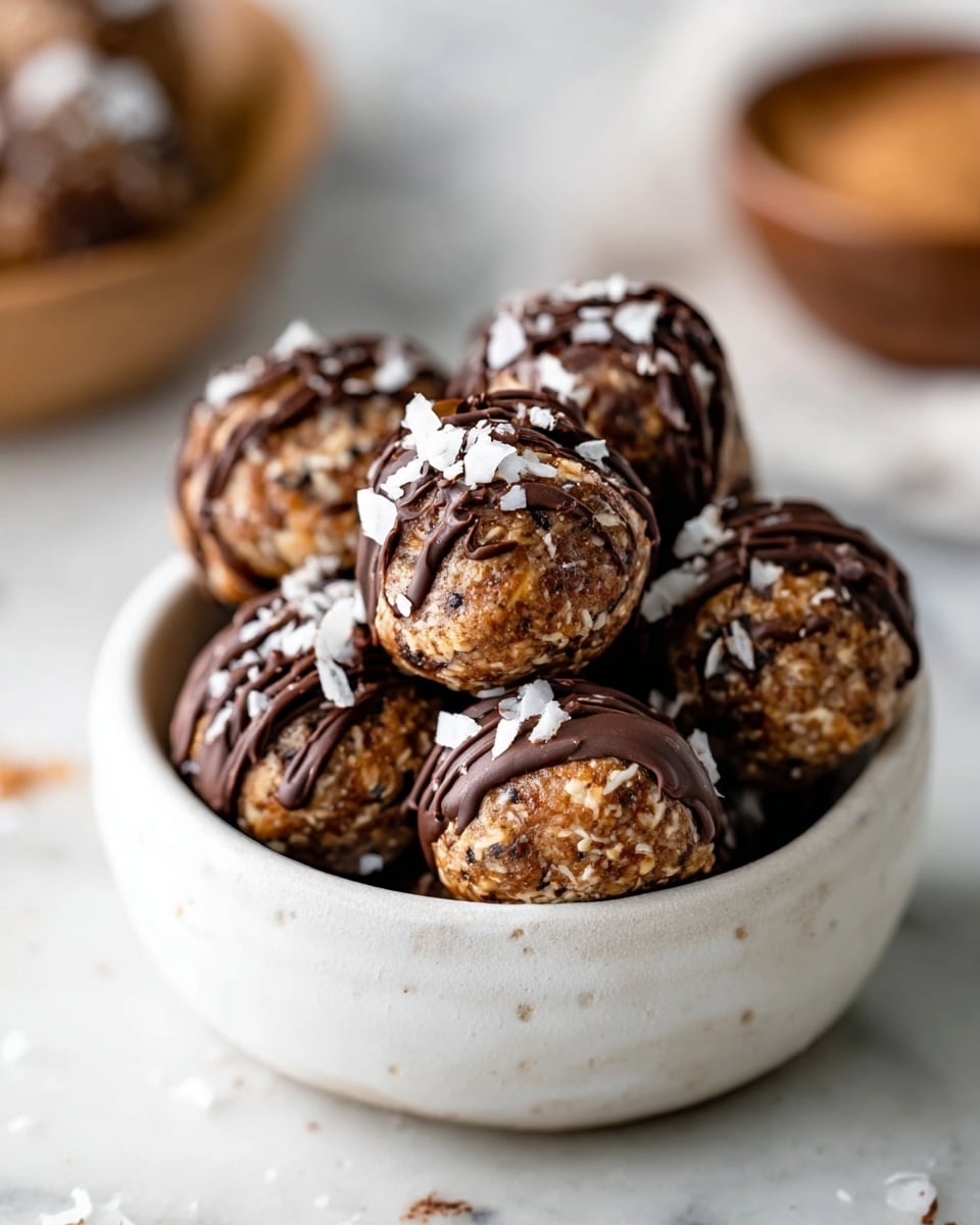 The image shows several round dessert balls arranged on a fluted metal tray, each with a golden brown base layer covered halfway in dark chocolate. The top half is drizzled with more dark chocolate and sprinkled with white coconut flakes, creating a textured contrast. One of the dessert balls sits in the front on a white marbled surface with scattered toasted coconut flakes and small dark chocolate chips. A small white plate filled with loose dark chocolate chips rests to the left side, and a soft beige cloth is seen in the blurred background. The lighting highlights the glossy chocolate and the rough texture of the coconut flakes. Photo taken with an iphone --ar 4:5 --v 7
