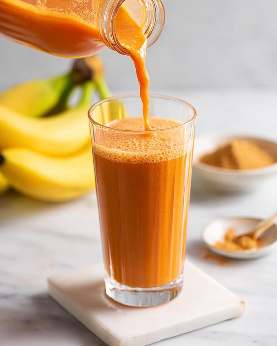 A clear glass bottle filled with smooth, thick orange carrot smoothie with a metal straw inside, placed on a small white marble square board on a white marbled surface. In the background, to the left, there are fresh carrots with green tops, and to the right, there are few yellow bananas beside another glass bottle containing the same orange smoothie. The overall setting is bright and clean. photo taken with an iphone --ar 4:5 --v 7