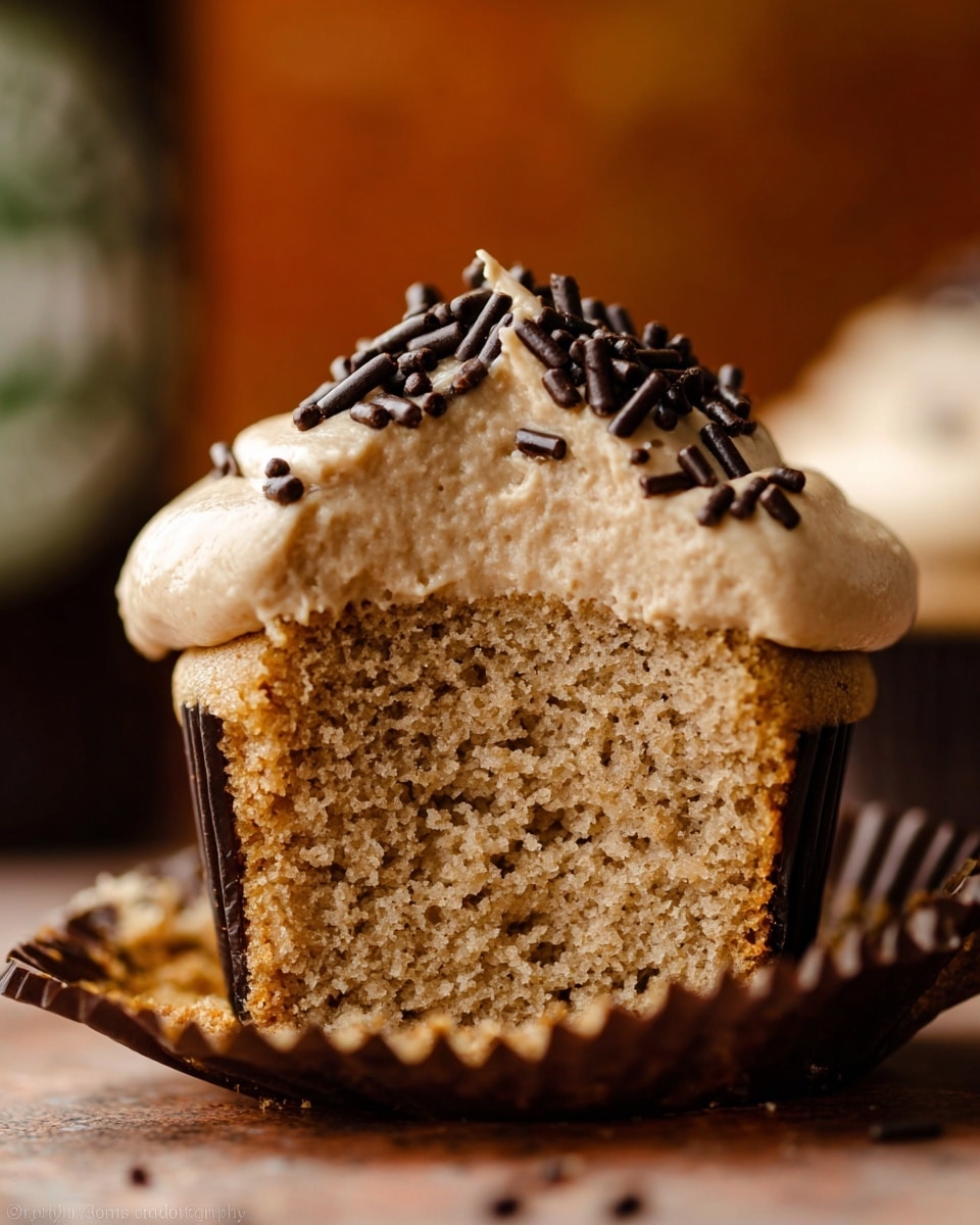 The image shows a close-up of a cupcake cut in half, revealing two layers. The bottom layer is a soft, light brown cake with a porous texture, while the top layer is a thick, smooth, light tan frosting. The frosting is decorated on top with dark brown, small, cylindrical sprinkles. The cupcake sits on a dark brown cupcake liner, and the background is a soft blur with warm tones. The focus is sharp on the cupcake's textures. Photo taken with an iphone --ar 4:5 --v 7