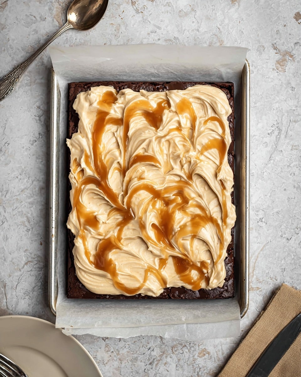 A square piece of dark brown chocolate brownie sits on a white speckled plate with a smooth layer of light beige frosting on top, swirled with a drizzle of caramel sauce creating a glossy amber swirl. A silver fork holds a bite-sized piece of the brownie with frosting, positioned on the left side of the plate. In the background, there is another white speckled plate with a partially eaten brownie and a fork, along with a box of Crisco shortening placed on a white marbled surface. photo taken with an iphone --ar 4:5 --v 7