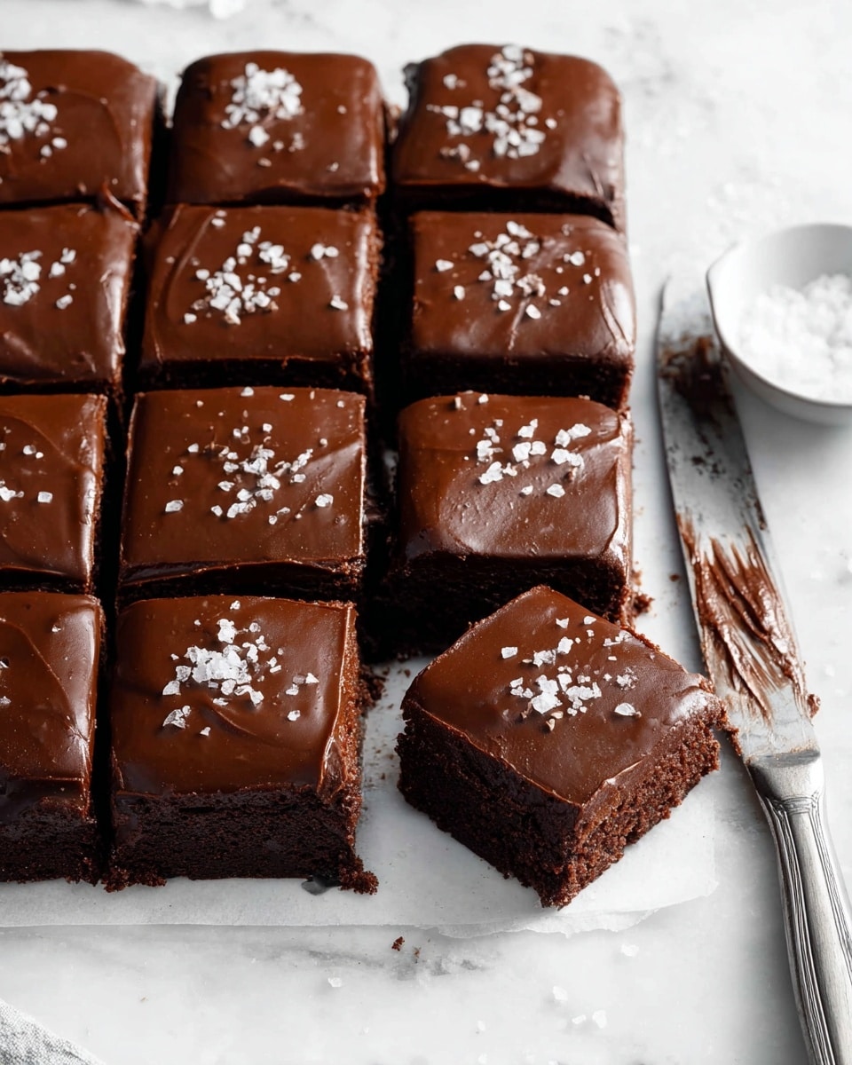 A clear glass bowl filled with smooth, thick chocolate batter of dark brown color, swirled with rich, creamy texture. Two metal beaters attached to an electric mixer are partially dipped into the batter, coated with the chocolate mixture, resting diagonally across the bowl. The bowl is placed on a white marbled surface, highlighting the glossy texture of the chocolate batter inside. photo taken with an iphone --ar 4:5 --v 7