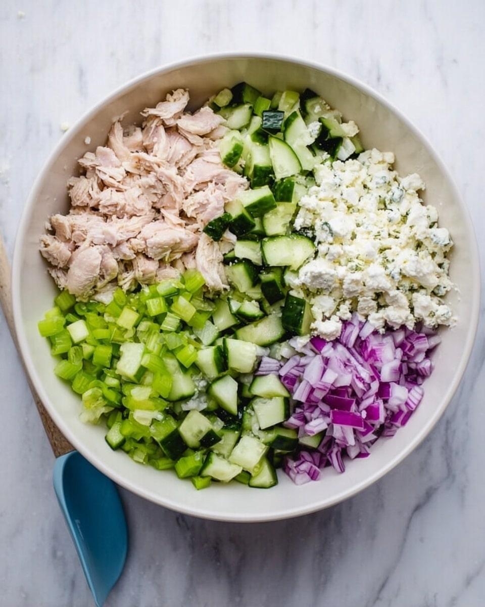A white bowl filled with a creamy salad that has multiple layers of colors and textures; the base layer is pale with small chunks of white and light green, mixed with finely chopped darker green herbs and small bits of purple onion scattered throughout. On top of the salad, two triangular brown crackers stand upright, partially inserted in the salad on the right side of the bowl. A silver spoon with a wooden handle lies inside the bowl, partially submerged in the salad near the bottom right. Around the bowl, more triangular brown crackers are placed on a white marbled surface, with a few green parsley leaves visible in the top right corner. photo taken with an iphone --ar 4:5 --v 7